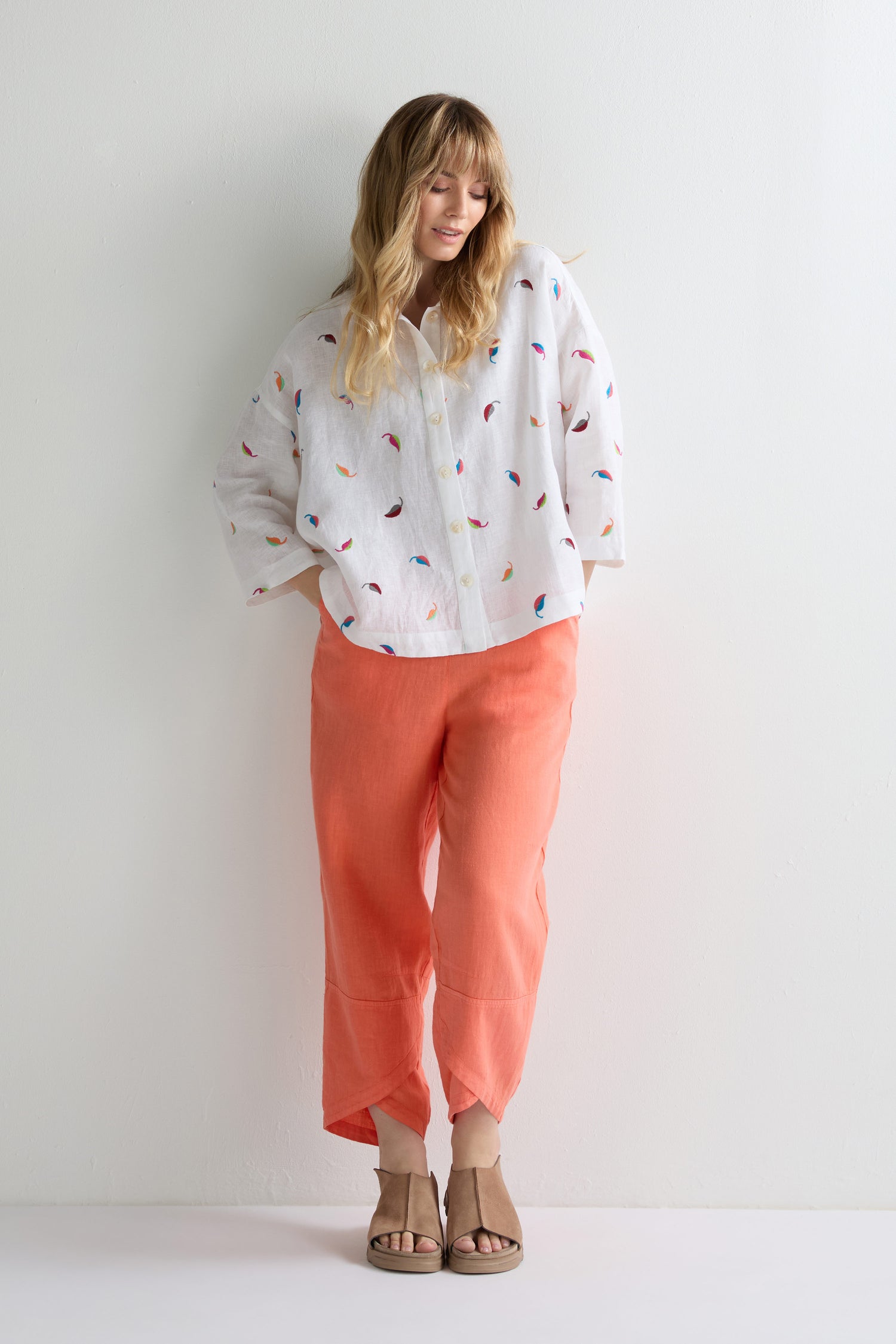 A woman stands against a white wall wearing the Embroidered Leaf Boxy Shirt, which features a colorful scattered pattern, paired with coral pants and brown sandals.