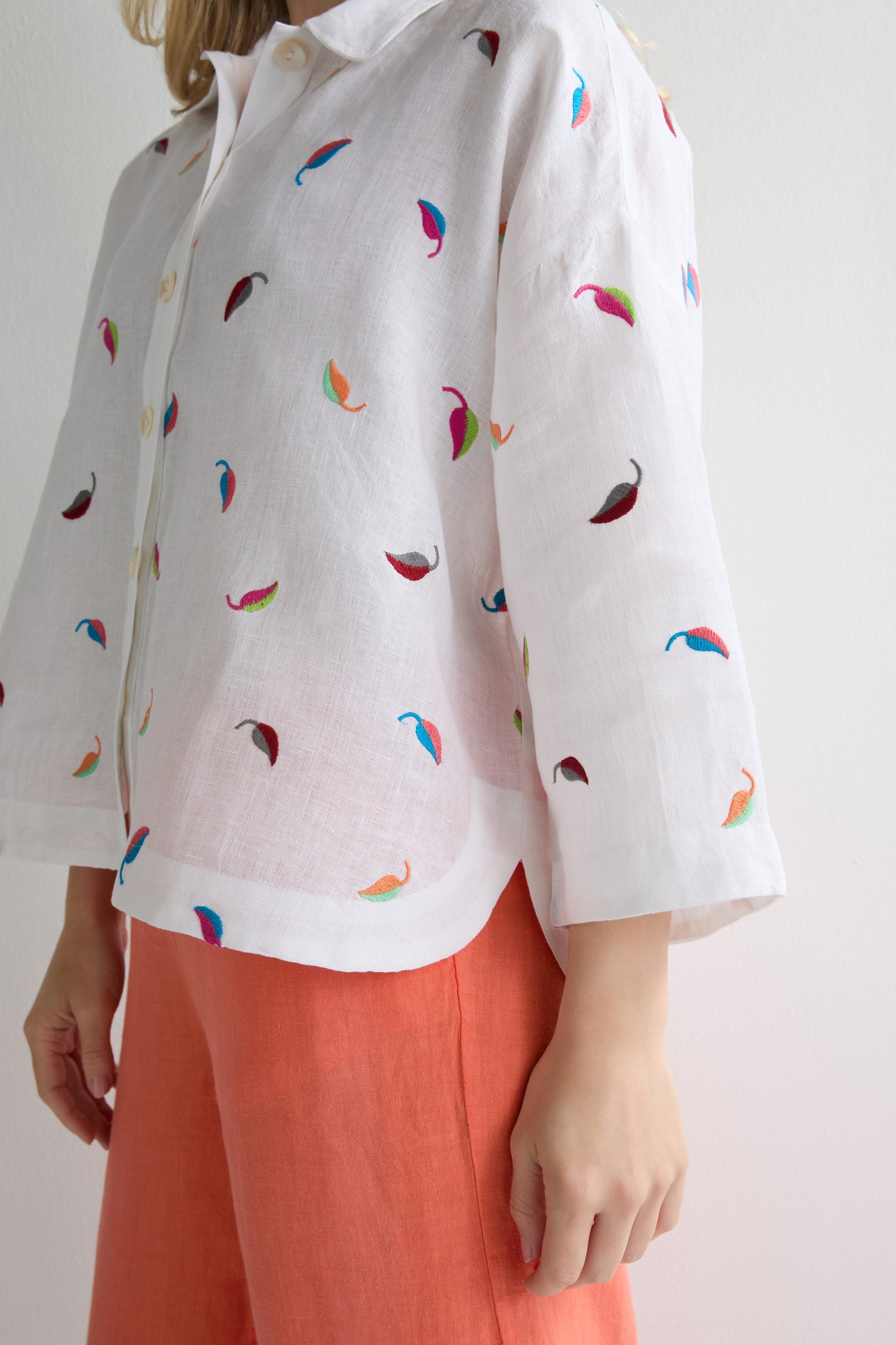 A person wearing the Embroidered Leaf Boxy Shirt in white, featuring colorful leaf embroidery, paired with coral-colored pants and standing against a plain light background.