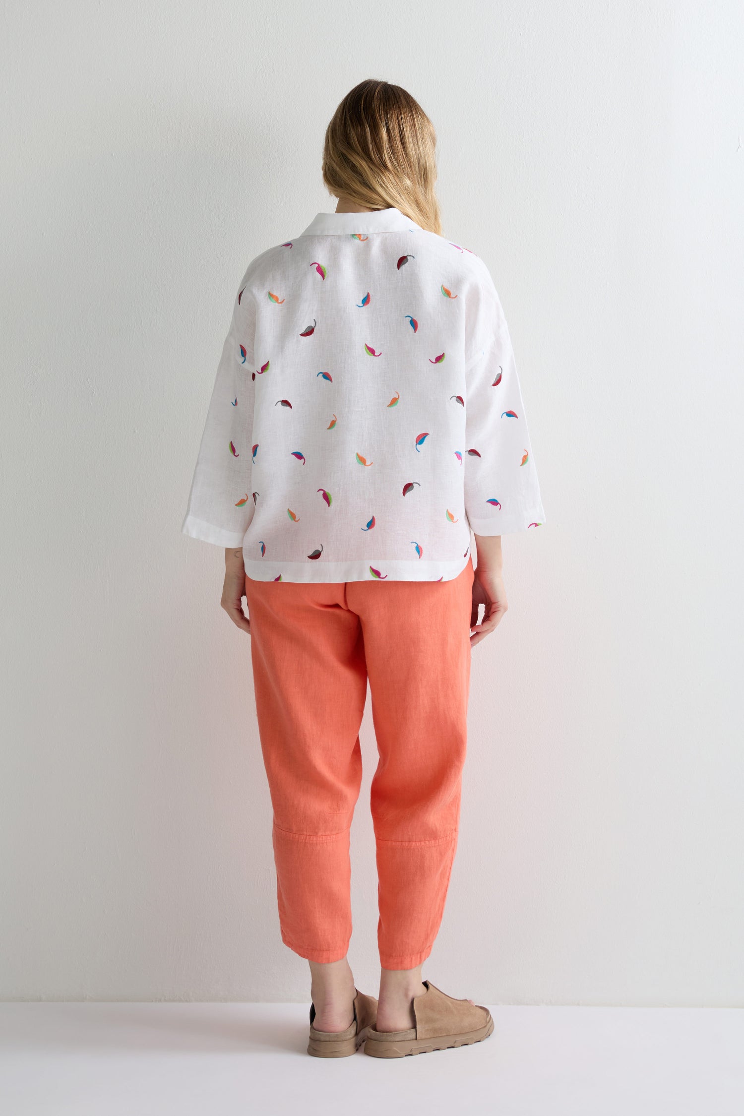 A person stands facing away, wearing the Embroidered Leaf Boxy Shirt with a colorful pattern, paired with coral pants and tan slip-on shoes against a plain light background.