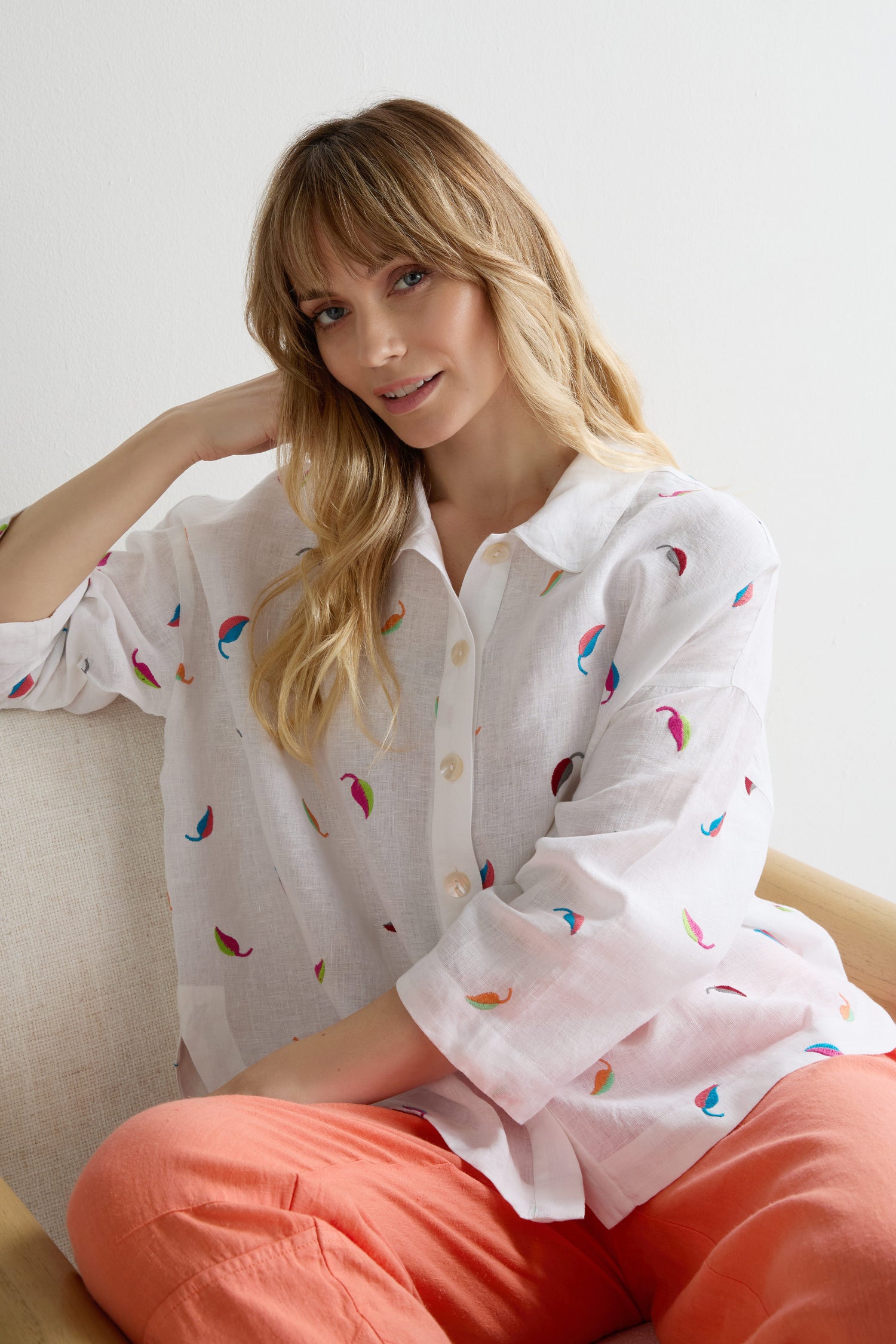 A woman with long blonde hair sits on a beige chair, wearing the Embroidered Leaf Boxy Shirt and coral pants, her left arm resting on the chair’s back.