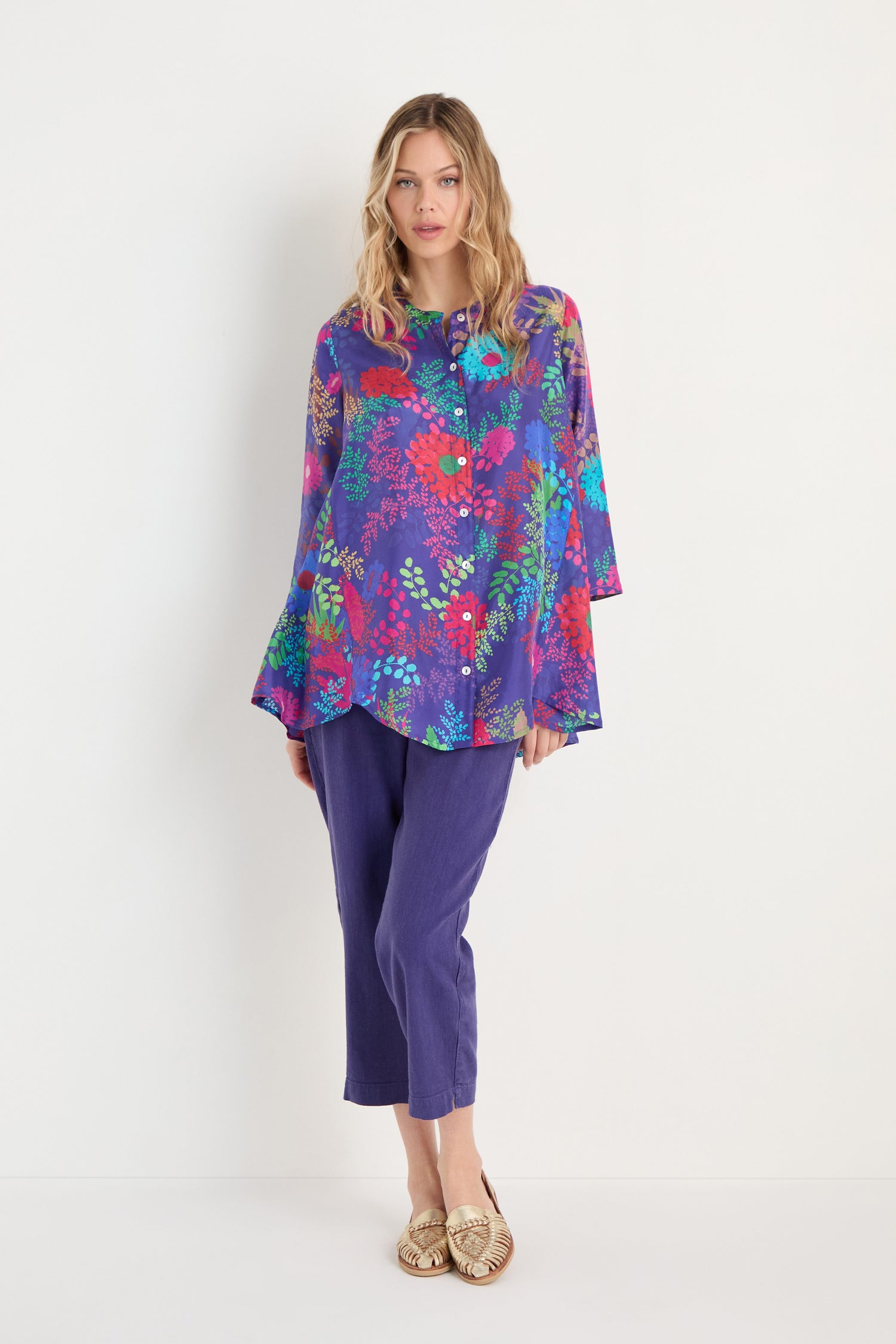 A woman stands against a white wall, dressed in the Bright Blooming Garden Silk Shirt with a botanical floral print, paired with purple cropped pants and gold flat shoes.