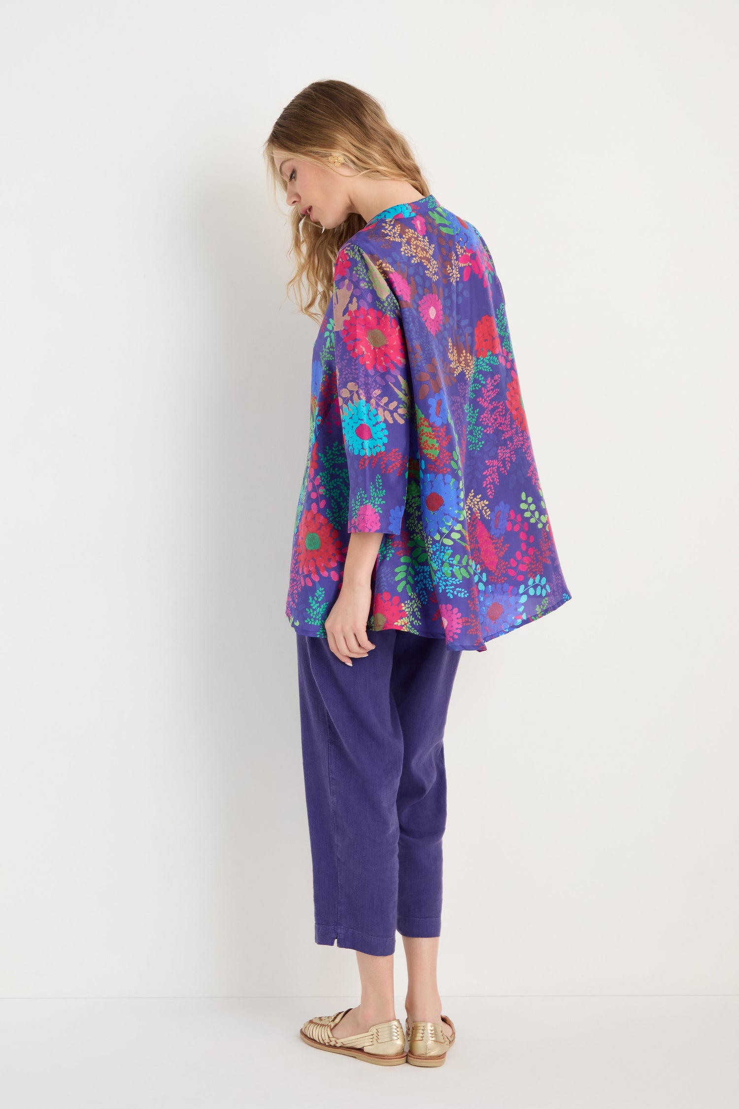 A woman stands sideways against a white background, wearing the Bright Blooming Garden Silk Shirt in a botanical purple floral print, paired with cropped purple pants and gold flats.