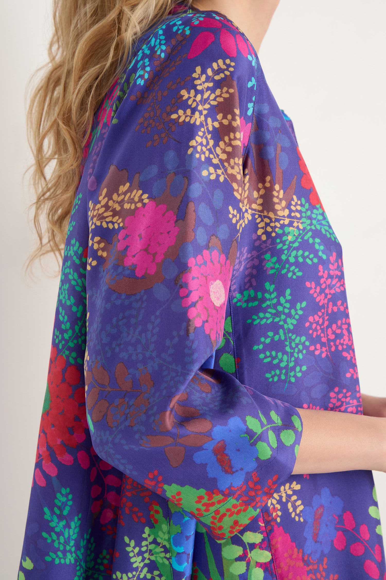 Someone wears the Bright Blooming Garden Silk Shirt, adorned with botanical-inspired floral patterns on a blue background, while standing against a plain, light-colored wall.