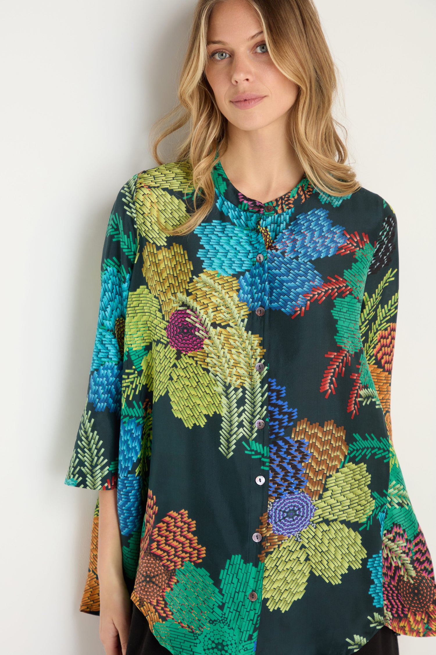 A woman with wavy blonde hair wears the Pixelated Petals Print Silk Shirt, a dark silk top with a pixelated floral design and front buttons.