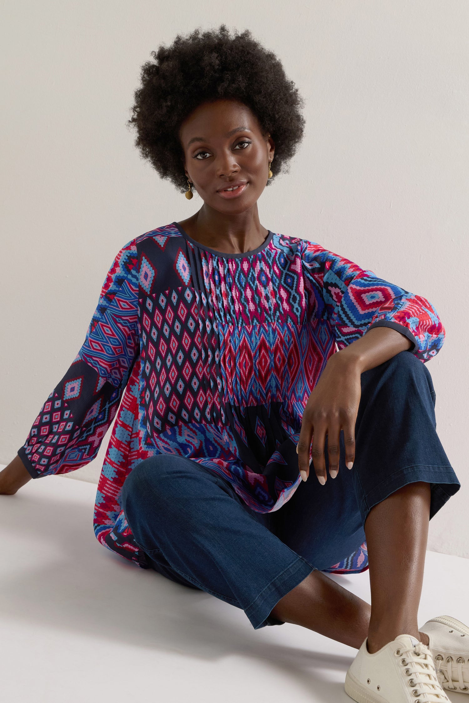 A person with an afro sits on the floor wearing a colorful Diamond Geo Patchwork Top, blue pants, and white sneakers.