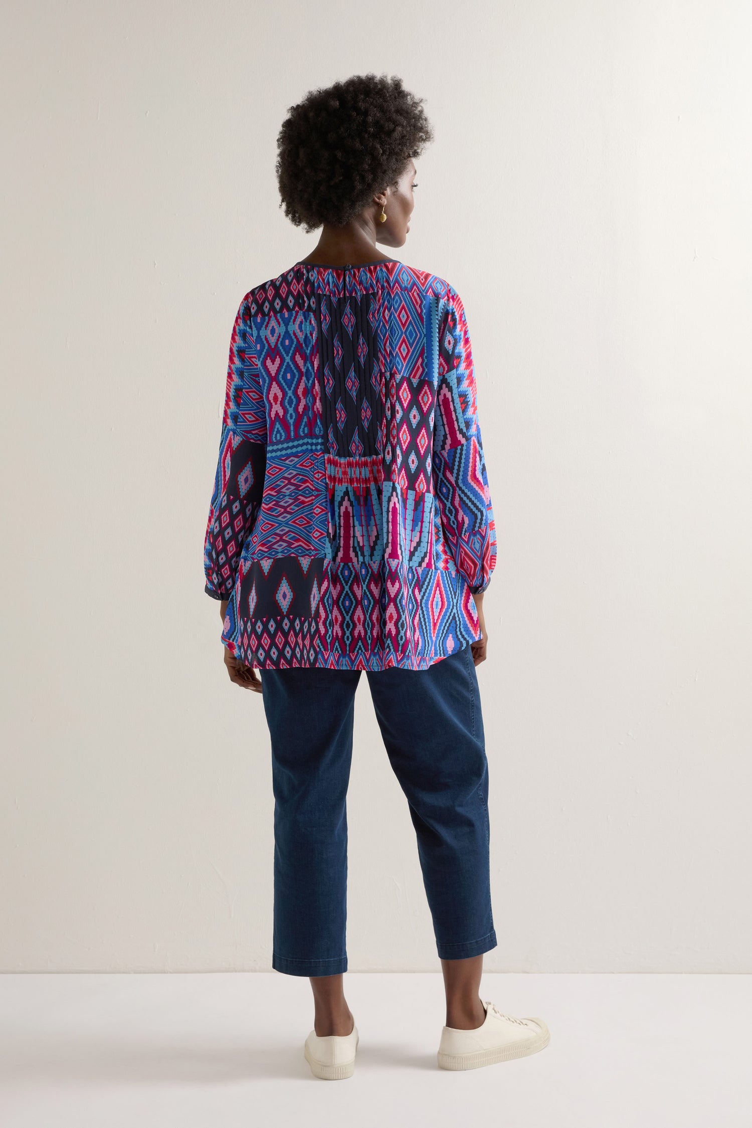 A person wearing a vibrant Diamond Geo Patchwork Top with dark jeans and white shoes stands back to the camera against a plain background.
