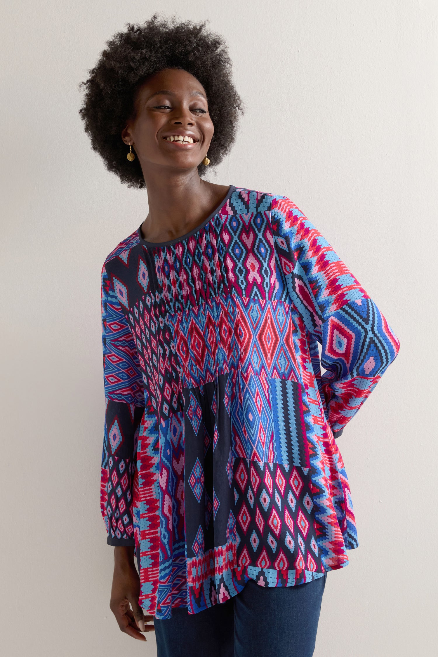 A person smiles against a plain background, wearing the Diamond Geo Patchwork Top with a colorful geometric pattern and jeans.