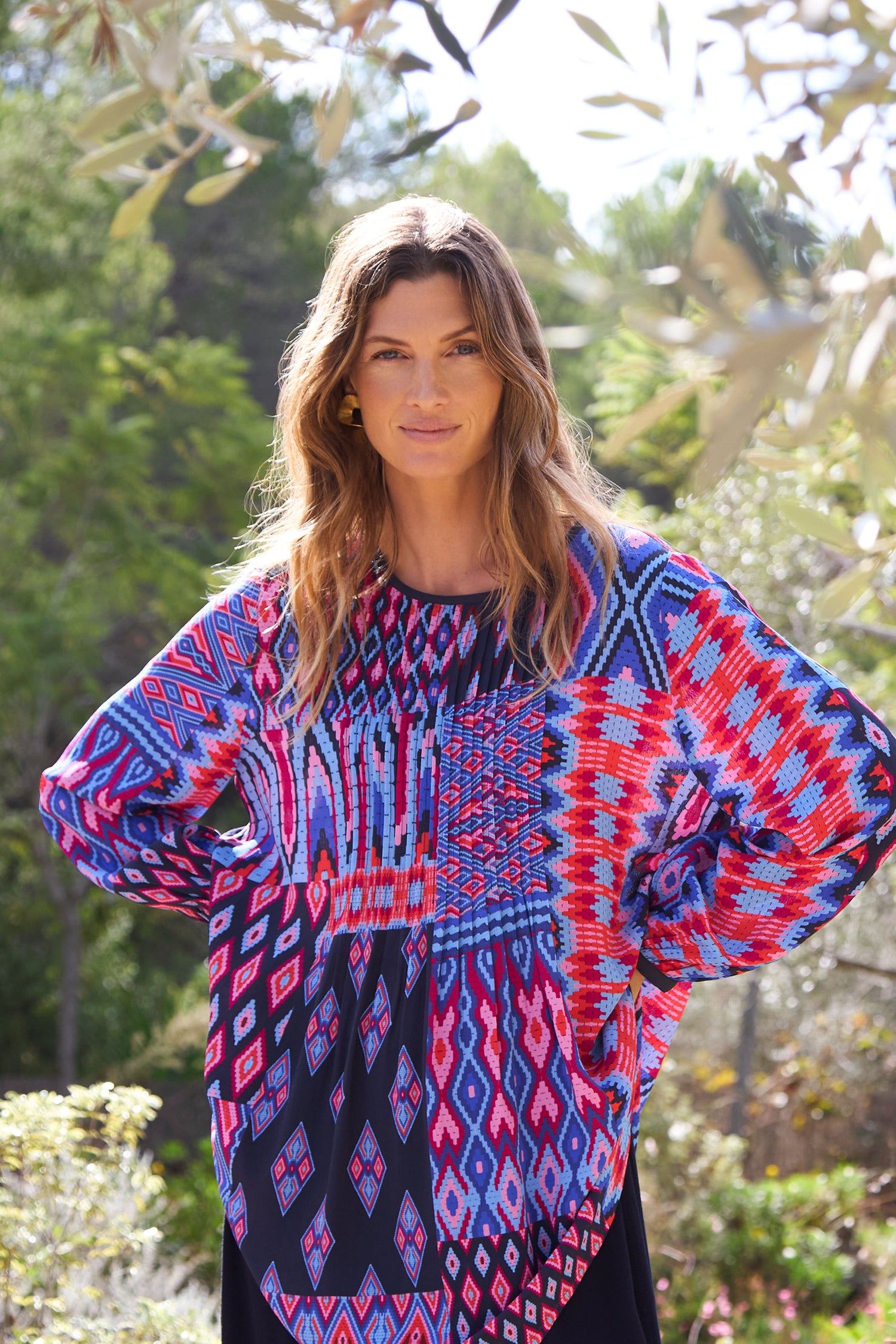 A woman stands amid lush greenery wearing a Diamond Geo Patchwork Top with a flattering fit-and-flare silhouette, enhancing her look in harmony with the vibrant landscape.