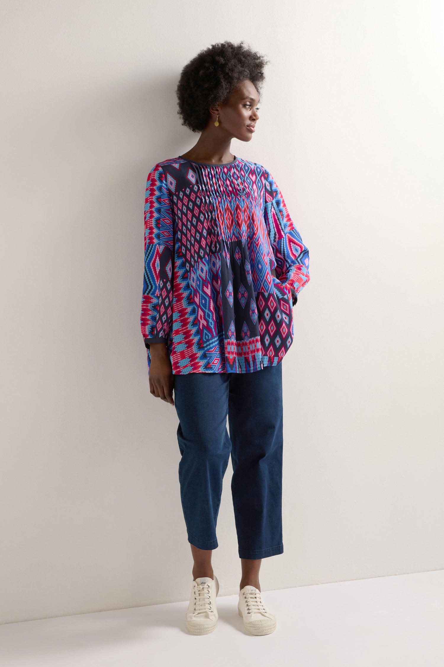 A person stands against a white wall, wearing the Diamond Geo Patchwork Top paired with blue jeans and white sneakers.