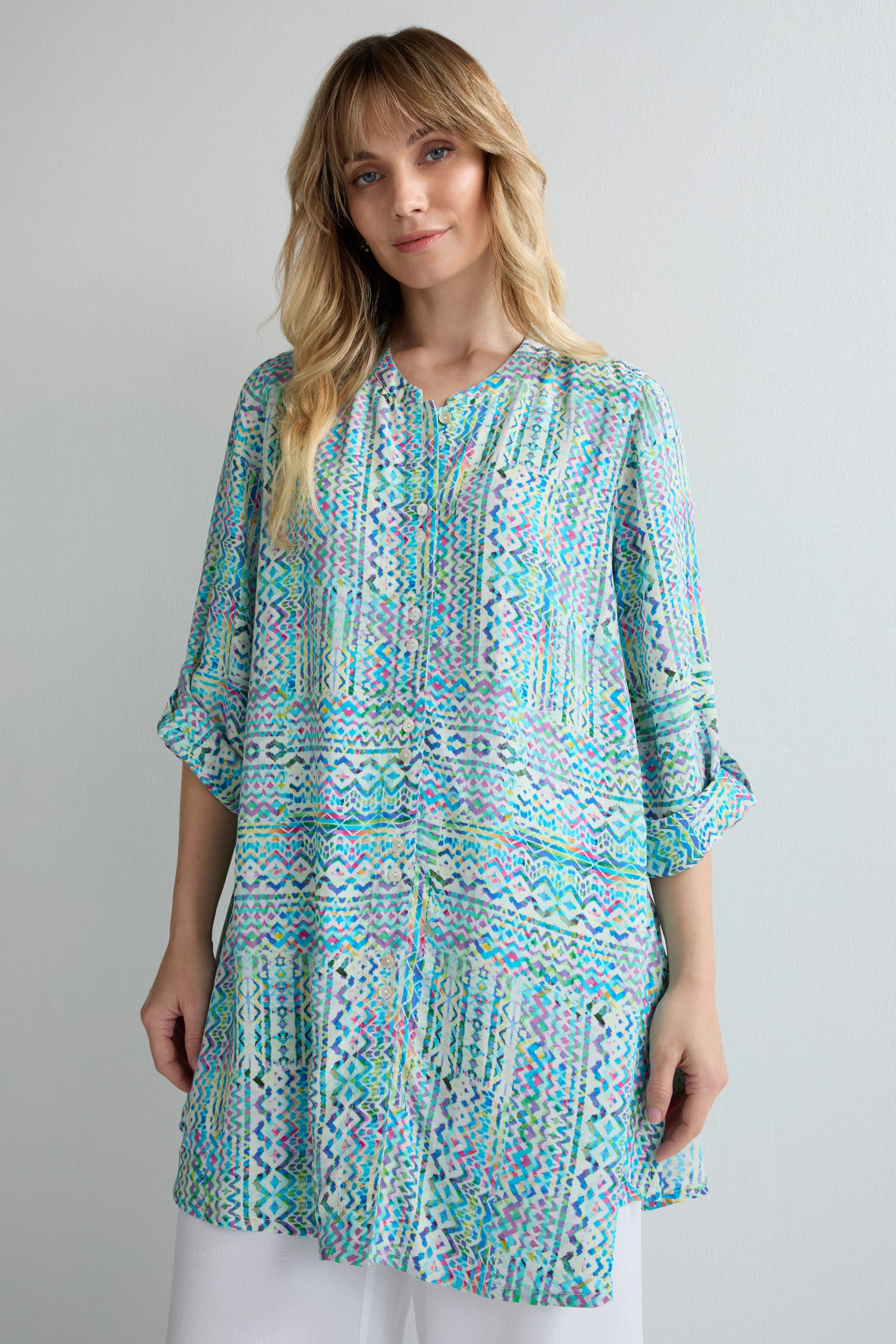 A woman stands against a plain light background wearing the Patchwork Tie Dye Shirt with rolled sleeves and white pants, highlighting its relaxed fit and colorful patchwork tie-dye design.