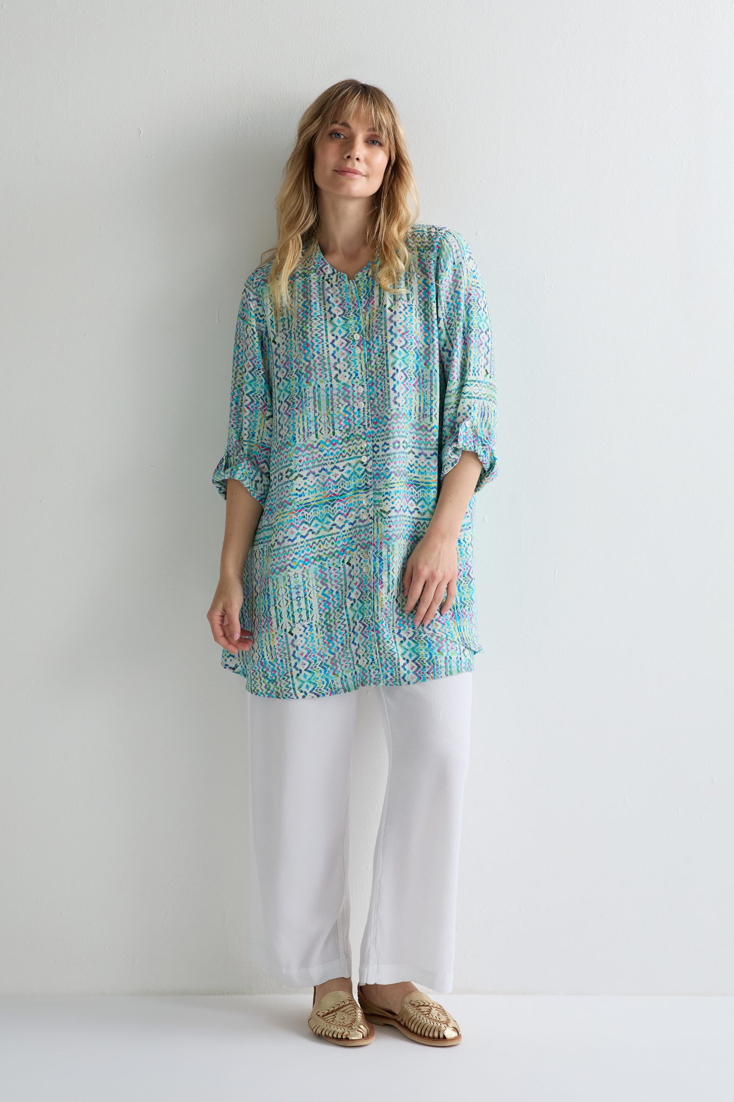A woman stands against a white wall wearing the Patchwork Tie Dye Shirt with relaxed tailoring, paired with white pants and gold flat sandals.