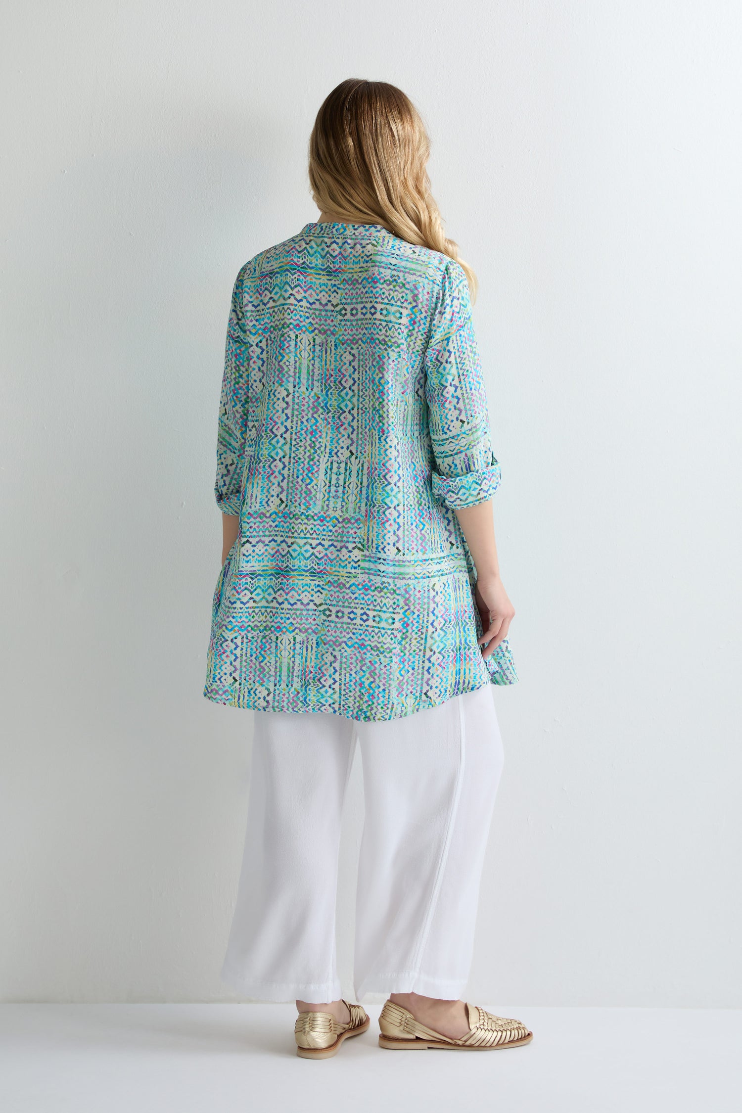 A person stands against a white wall, facing away, wearing a Patchwork Tie Dye Shirt in turquoise and blue, paired with white wide-leg pants and gold shoes.