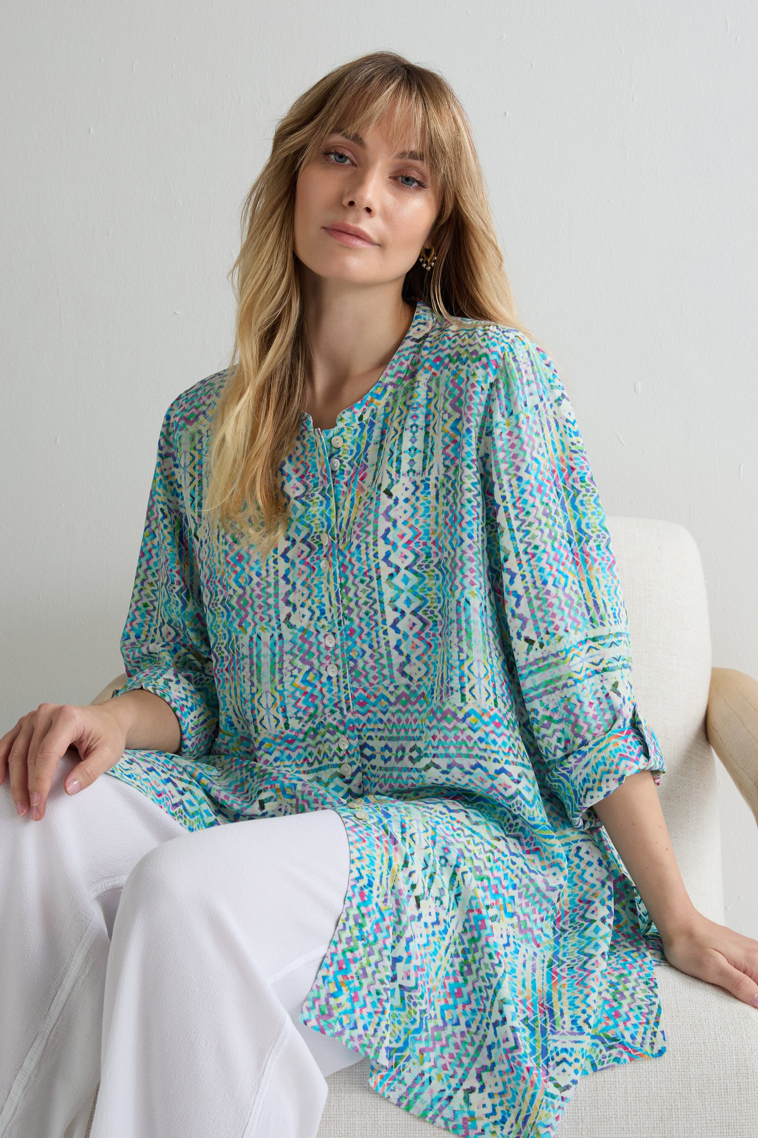 A woman with long blonde hair wears a vibrant Patchwork Tie Dye Shirt and white pants, sitting on a light-colored chair against a plain background.