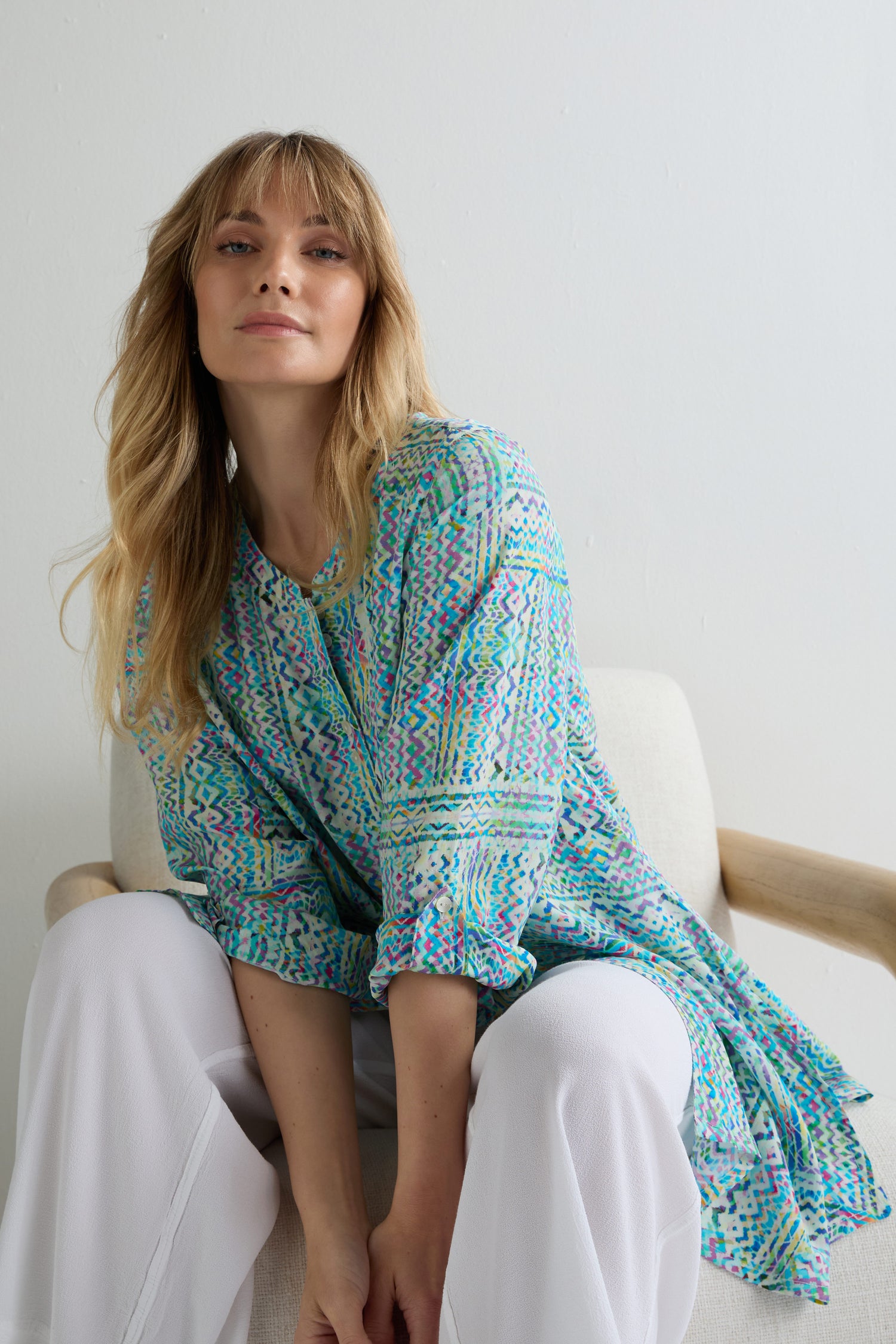 A woman with long blonde hair sits on a light chair, wearing the Patchwork Tie Dye Shirt with relaxed tailoring and white pants, looking slightly upward at the camera.