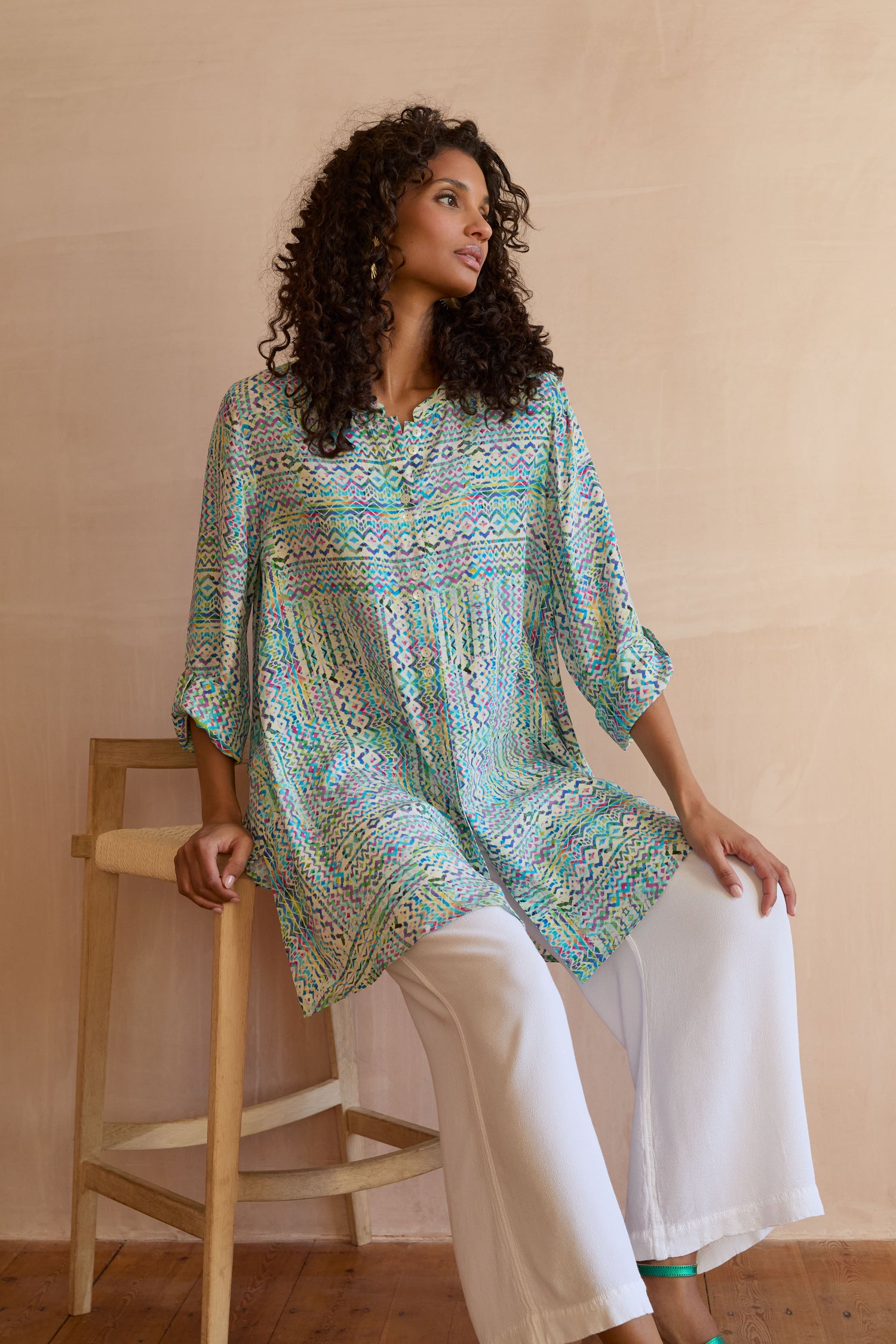 A woman with curly hair sits on a wooden stool, wearing the Patchwork Tie Dye Shirt in green and blue with white wide-leg pants, highlighting its relaxed fit against a neutral background.