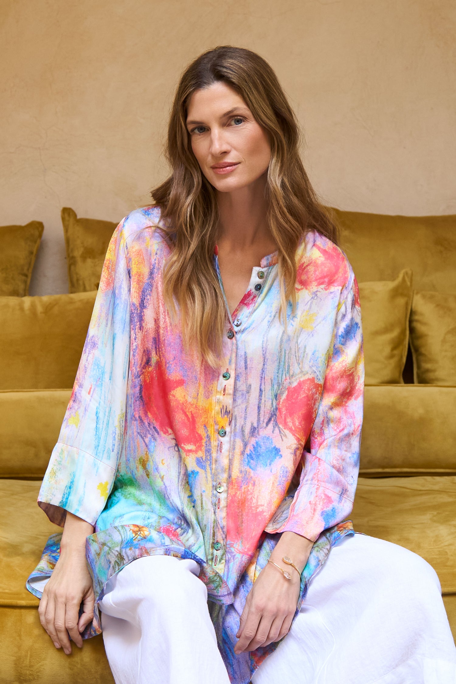 A woman with long brown hair sits on a yellow sofa, wearing the Meadow Dreams Flared Shirt and white pants, looking at the camera with a neutral expression.