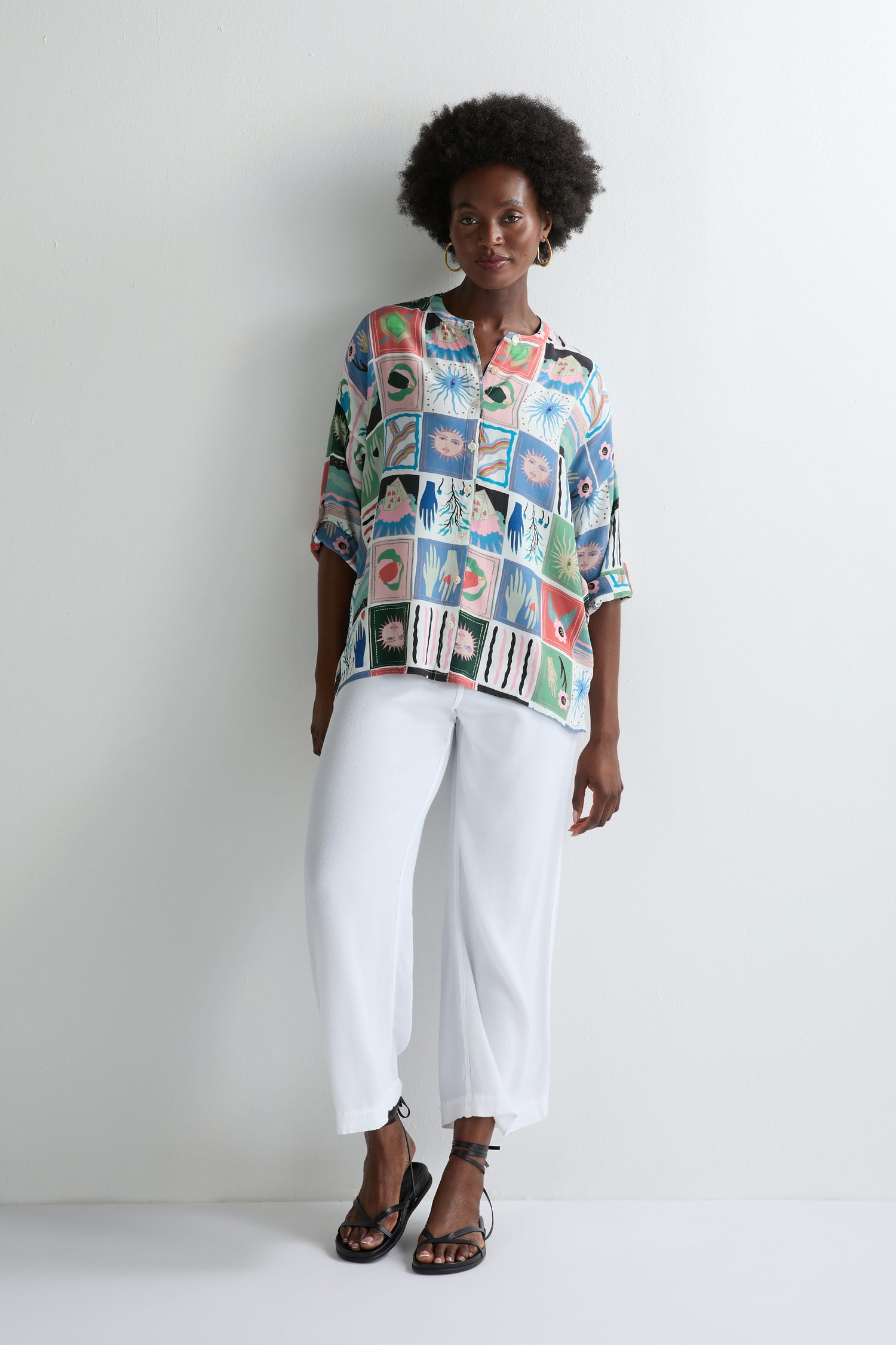 A person stands against a white wall wearing the Mystical Collage Boxy Shirt, styled with loose white pants and black sandals.