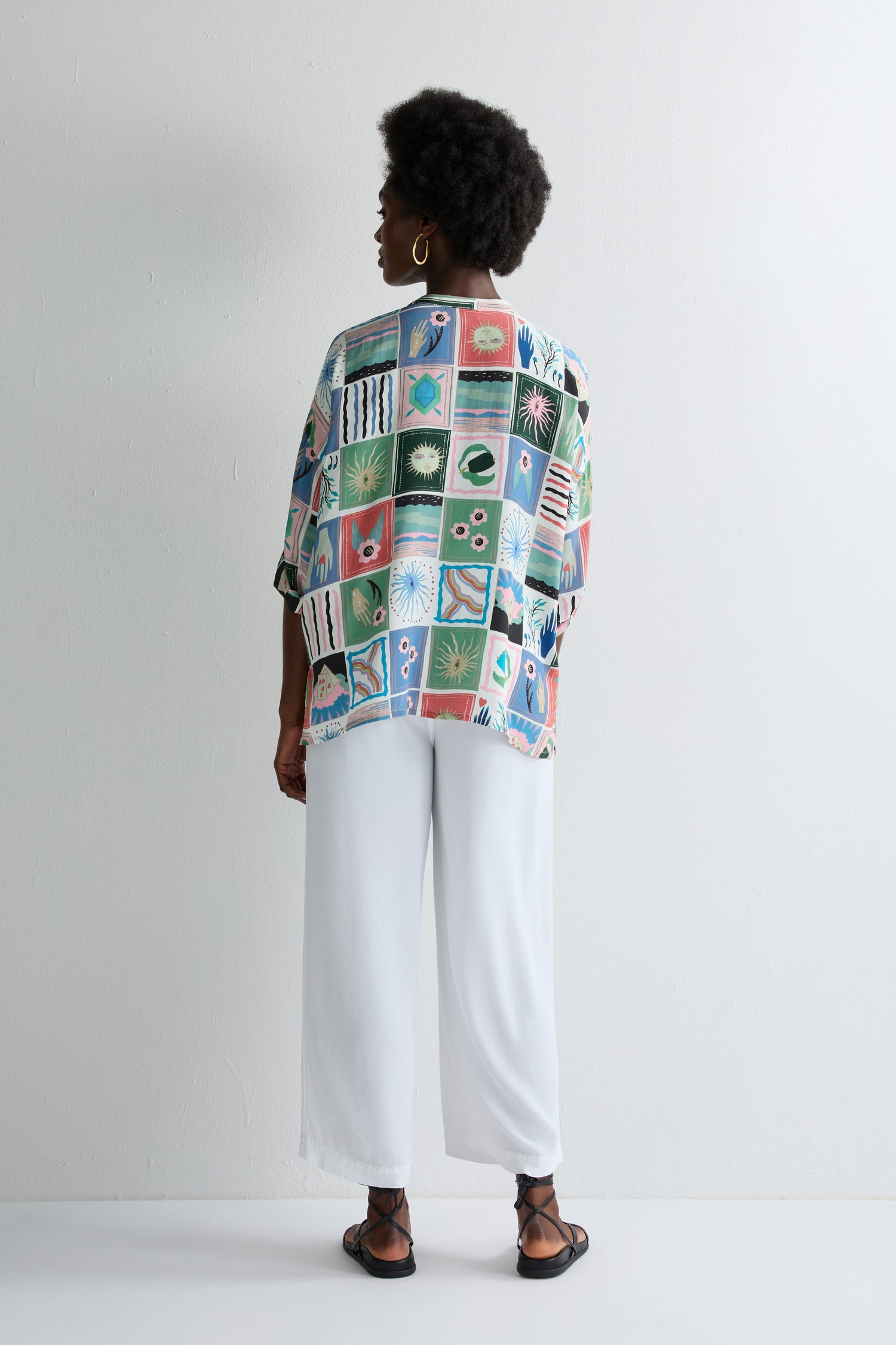 A person faces a white wall, wearing the Mystical Collage Boxy Shirt with a relaxed boxy fit and mandarin collar, styled with white pants, black sandals, and hoop earrings.