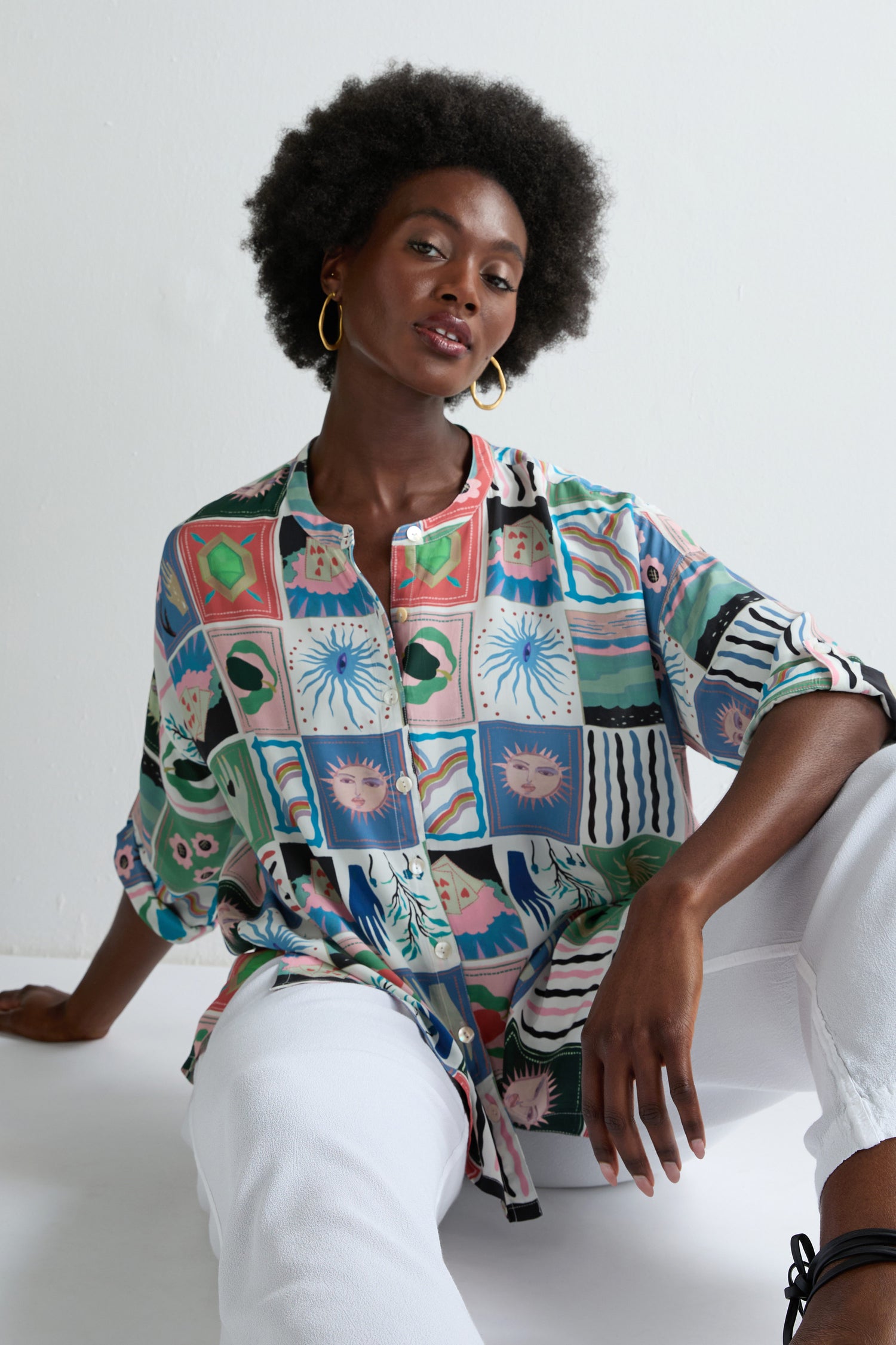 A woman with short curly hair sits on the floor against a plain white background, wearing the Mystical Collage Boxy Shirt and white pants.