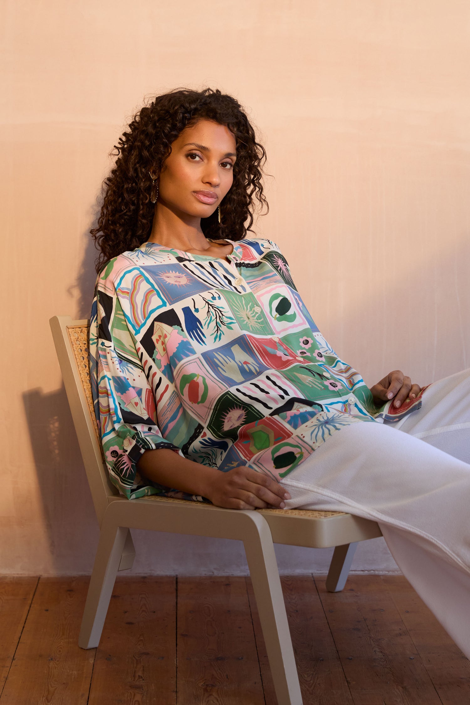 A woman with curly hair sits against a peach wall, wearing the Mystical Collage Boxy Shirt with a mandarin collar and white pants, highlighting its relaxed, boxy fit.