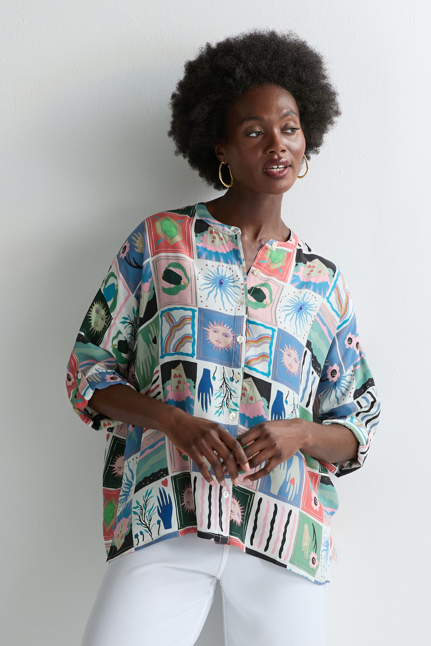 A woman with short curly hair stands against a white wall, wearing the Mystical Collage Boxy Shirt with a nature motif and mandarin collar. She pairs it with white pants and gazes to the side, creating an effortlessly stylish look.