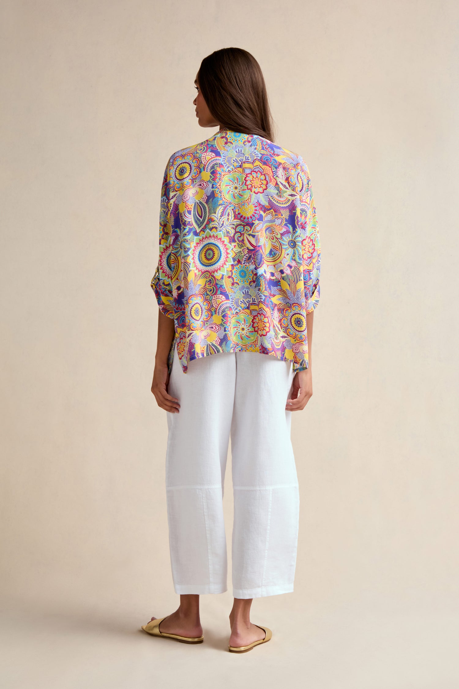 A woman stands facing away, wearing the Summer Sarasa Relaxed Shirt with a boxy silhouette, white wide-leg pants, and beige flat shoes against a plain beige background.