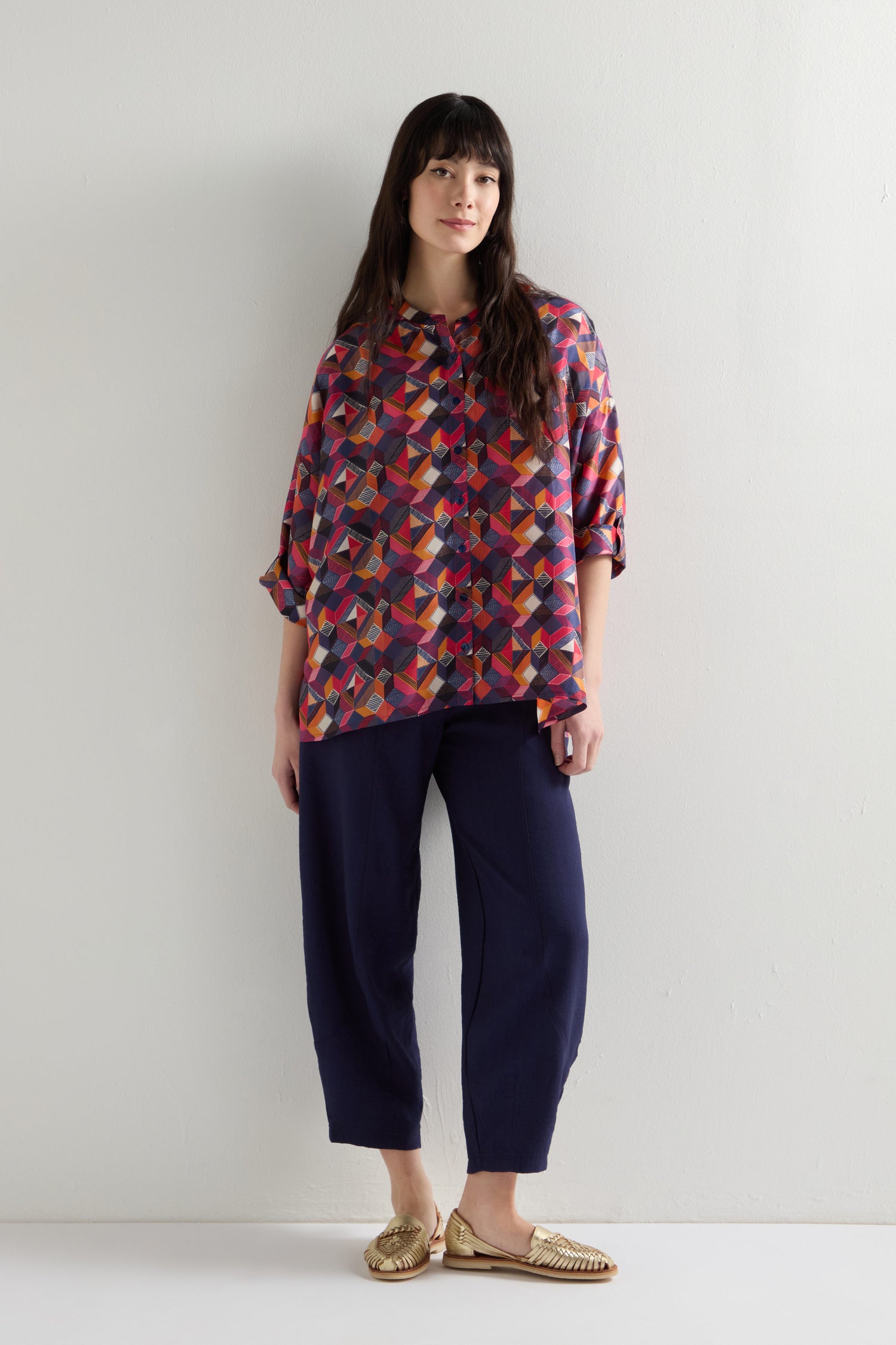 A person stands against a plain background wearing an oversized Small Stitched Squares Silk Shirt, dark pants, and gold shoes.