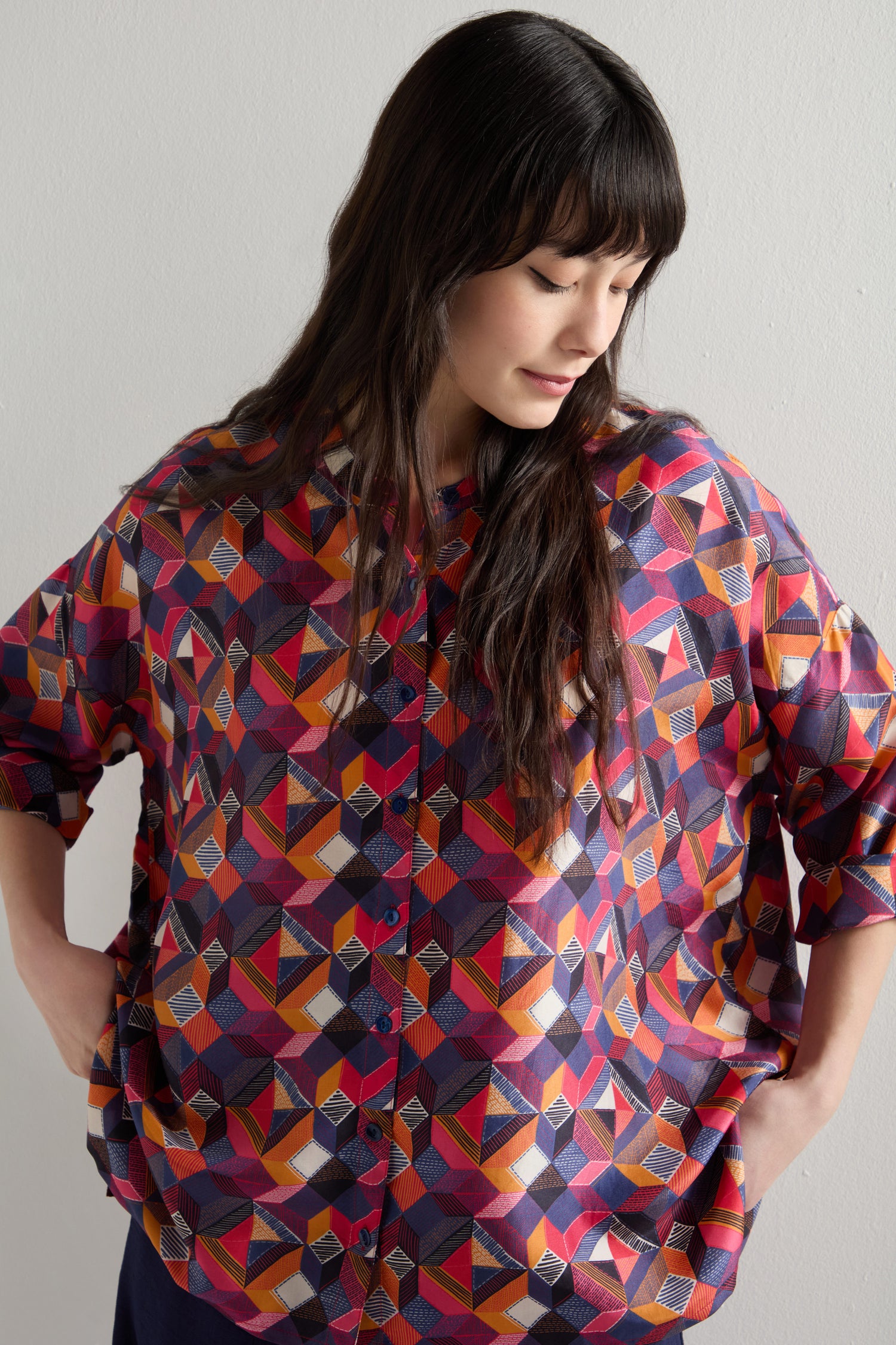 A person, standing with hands in pockets and gazing downward, wears an oversized Small Stitched Squares Silk Shirt featuring a vibrant geometric square print.