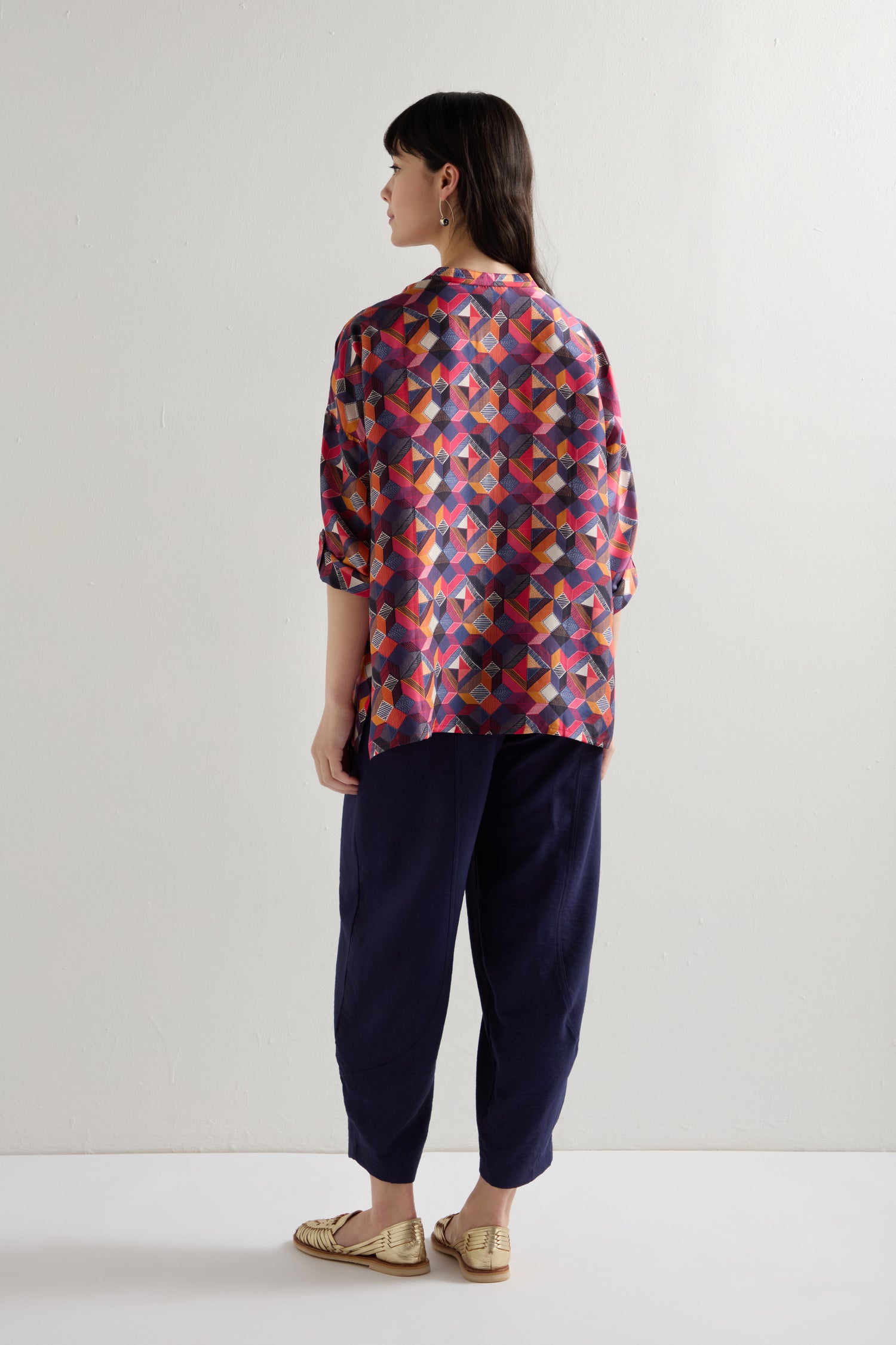 Facing away, a person wears an oversized fit "Small Stitched Squares" silk shirt with a colorful geometric square print, navy pants, and gold shoes against a plain white background.
