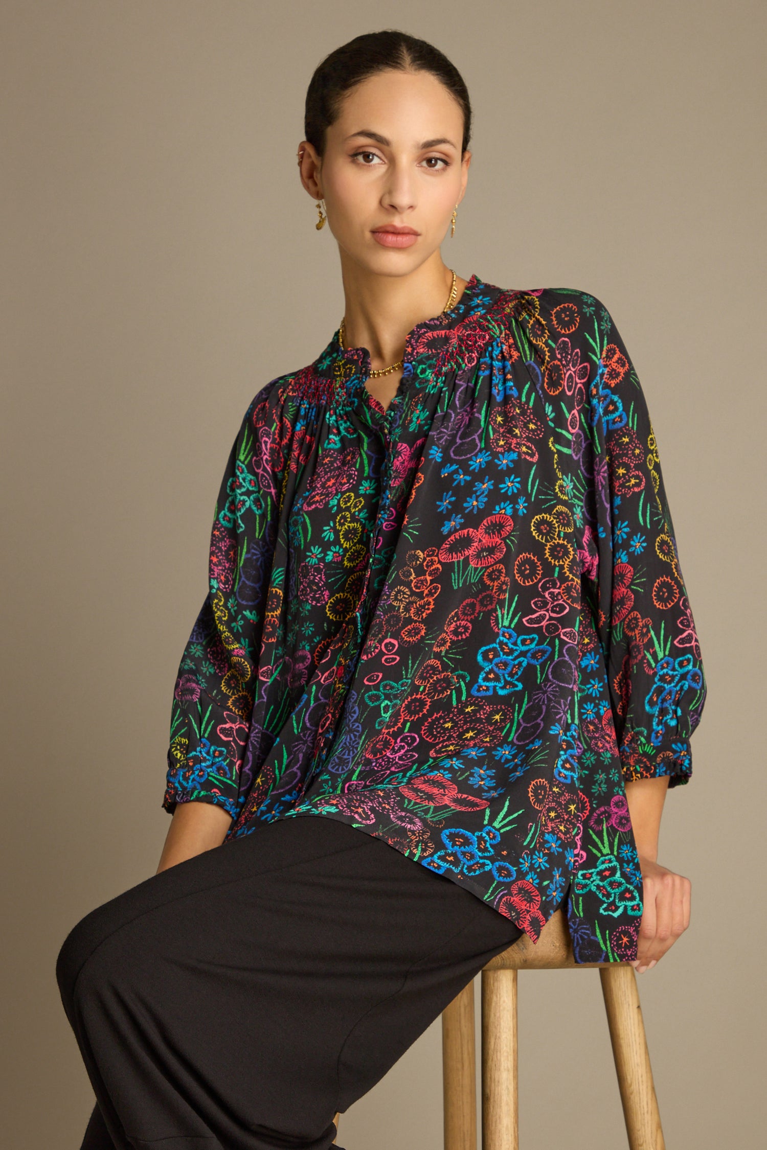 A person sits on a wooden stool wearing a Floral Embroidery Print Shirt and black pants, set against a neutral background.