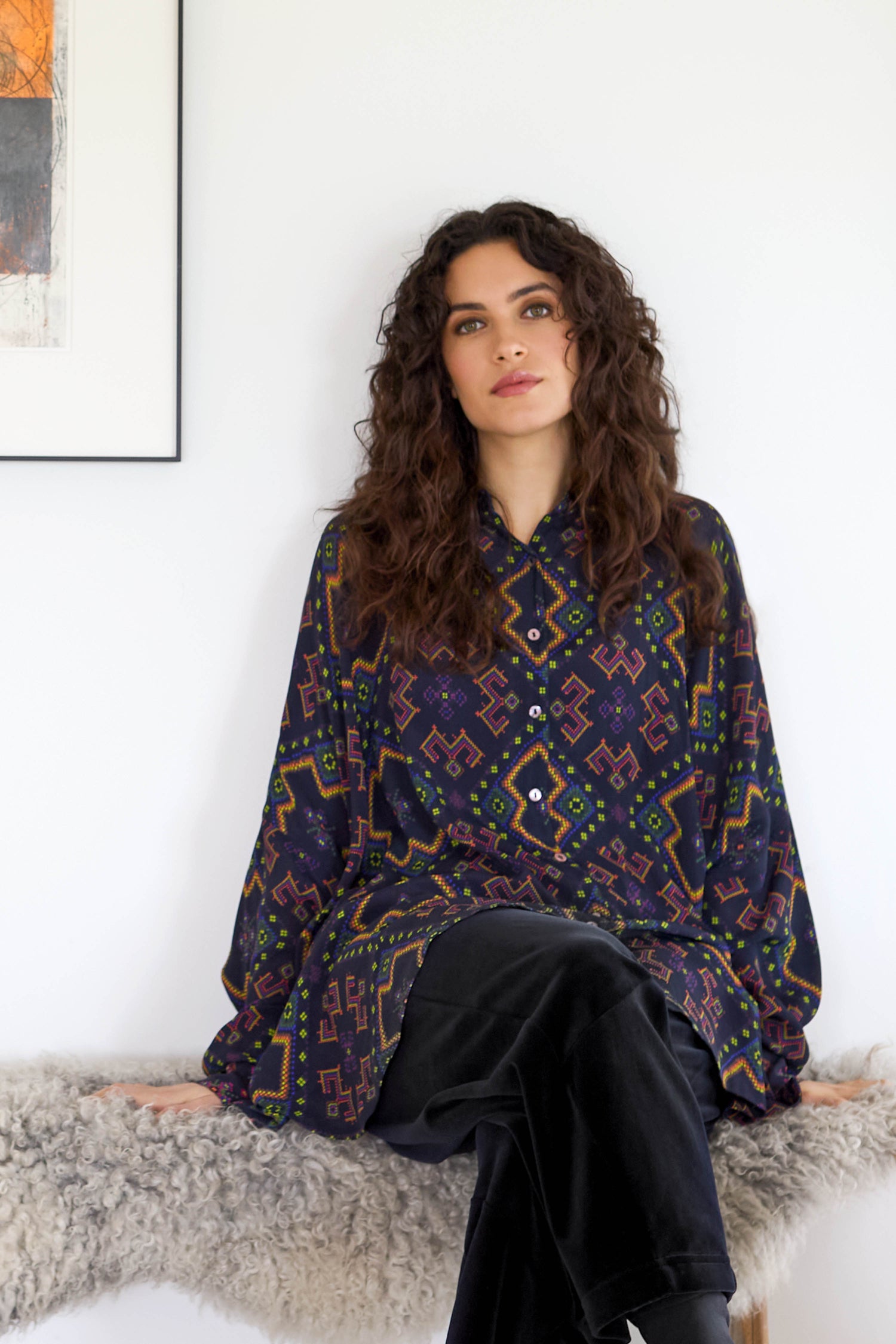 A woman with long curly hair sits on a bench, wearing a Pixelated Jacquard Print Shirt made of silky crepe viscose and black pants. A framed artwork is on the wall behind her.