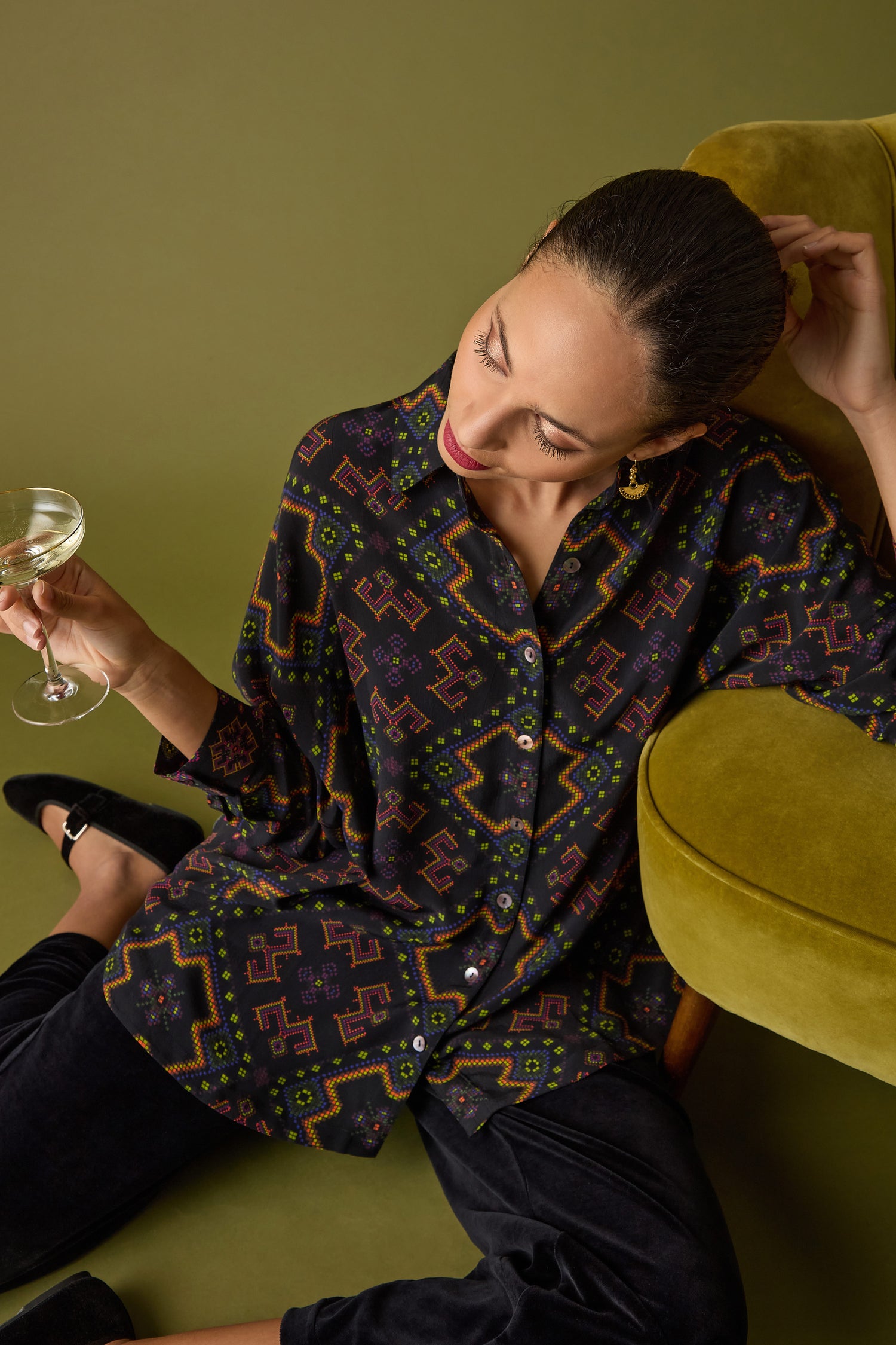 A person sits on the floor, wearing a Pixelated Jacquard Print Shirt, holding a glass of white wine while leaning against a green chair.
