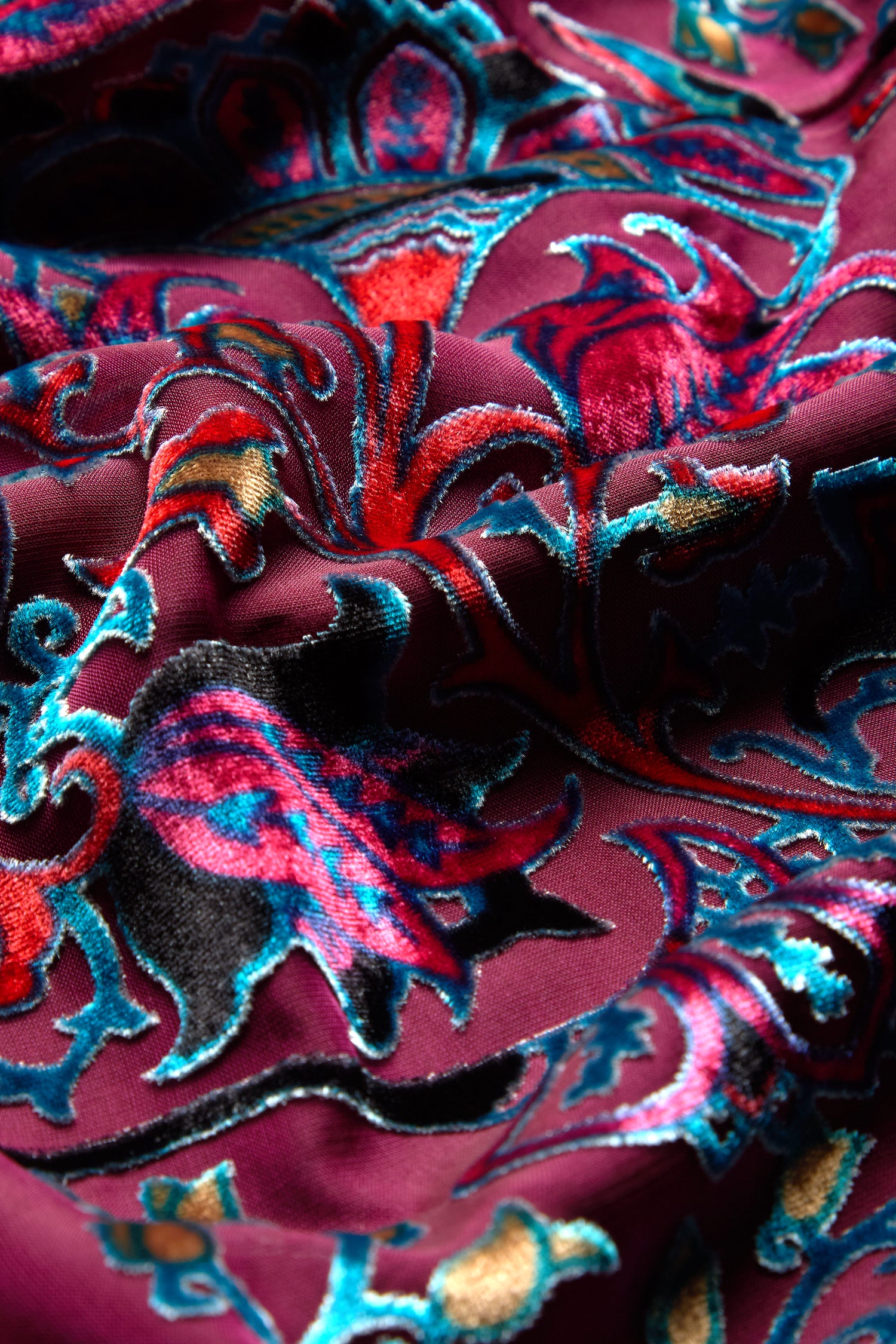 Close-up of a fabric adorned with complex pink, blue, and gold floral patterns set against a maroon background, reminiscent of the design found on the Winter Paisley Devore Top.