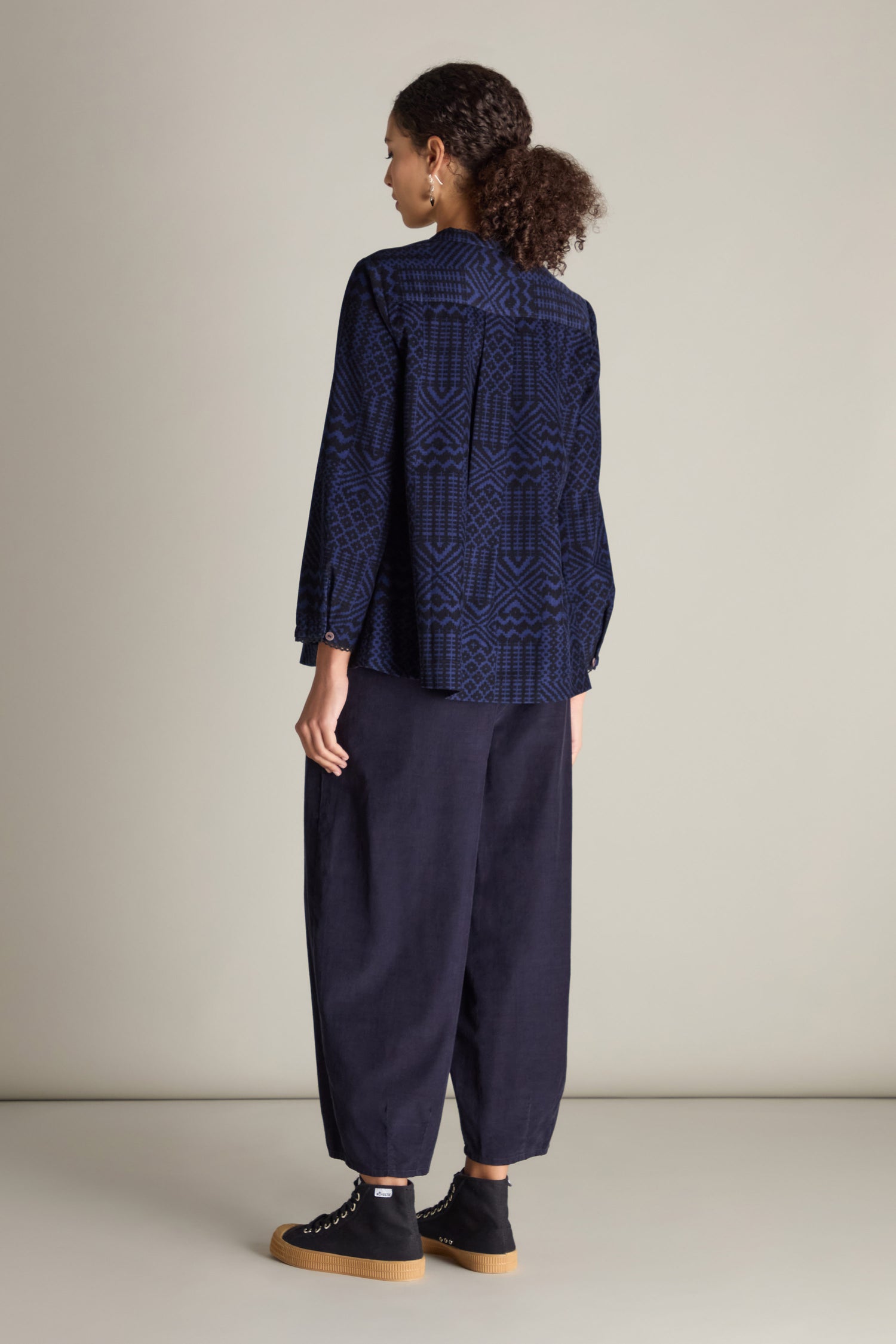 A person stands with their back to the camera, wearing a Mono Ikat Printed Cord Shirt along with loose navy pants and black sneakers featuring white soles. The ikat-inspired print of the shirt lends an elegant touch. They have curly hair tied back, and the background is plain and light-colored.