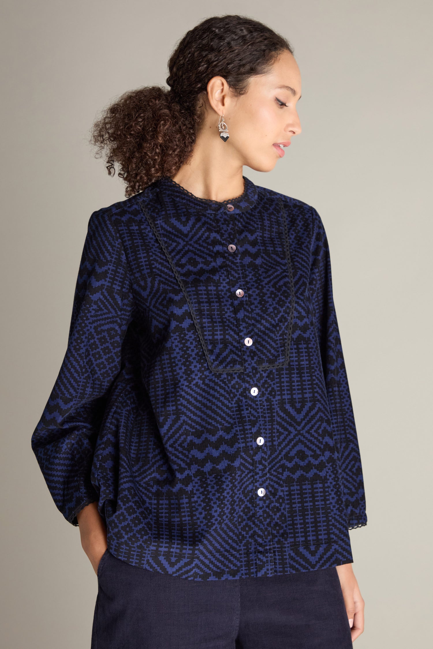 A woman with curly hair is wearing a Mono Ikat Printed Cord Shirt in dark blue with long sleeves, and she is looking to the side against a plain background.