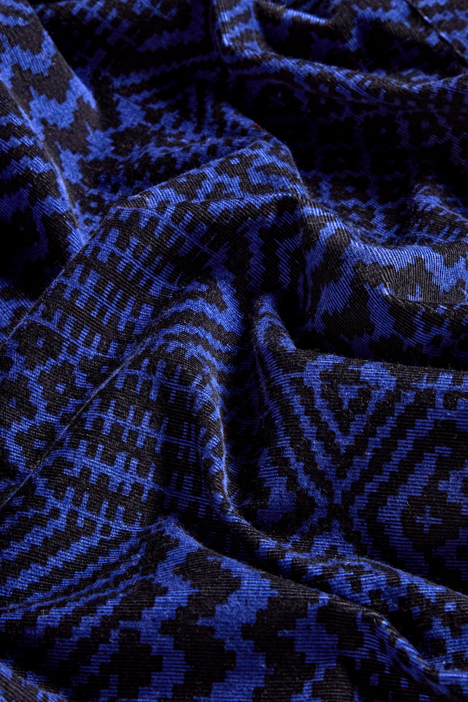A detailed view of the Mono Ikat Printed Cord Shirt, displaying its blue and black fabric with an intricate Ikat-inspired geometric pattern. The texture reveals folds and creases in the material, adding depth to its striking design.
