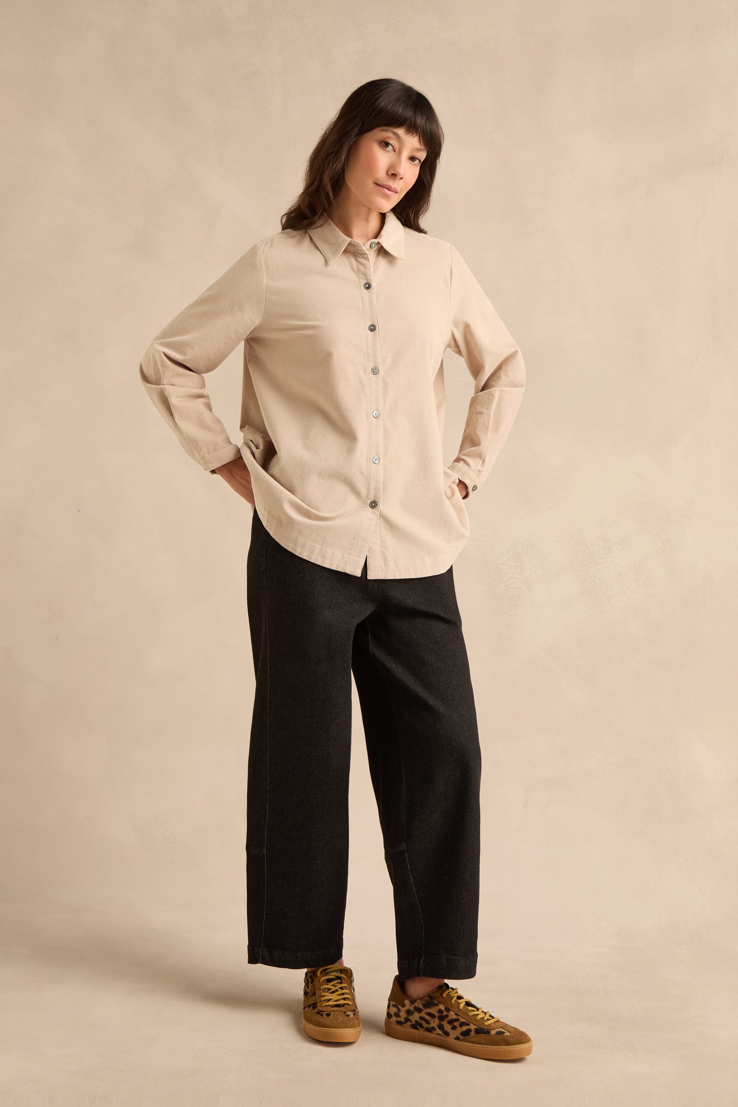 A person stands against a beige background, wearing the Pinwale Cord Shirt in pure cotton beige, black wide-leg pants, and leopard print sneakers.