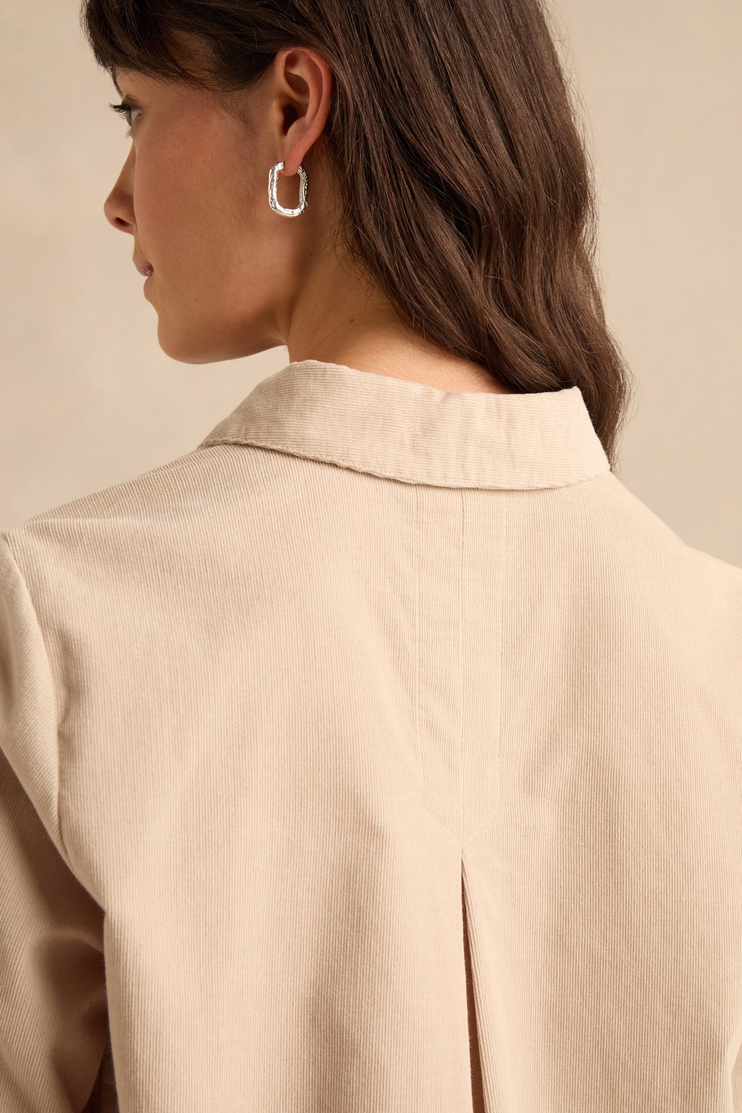 A woman with long brown hair wears the Pinwale Cord Shirt in beige, featuring a pure cotton fabric and a collared neckline, paired with a silver hoop earring; she is shown from behind and in profile against a neutral background.