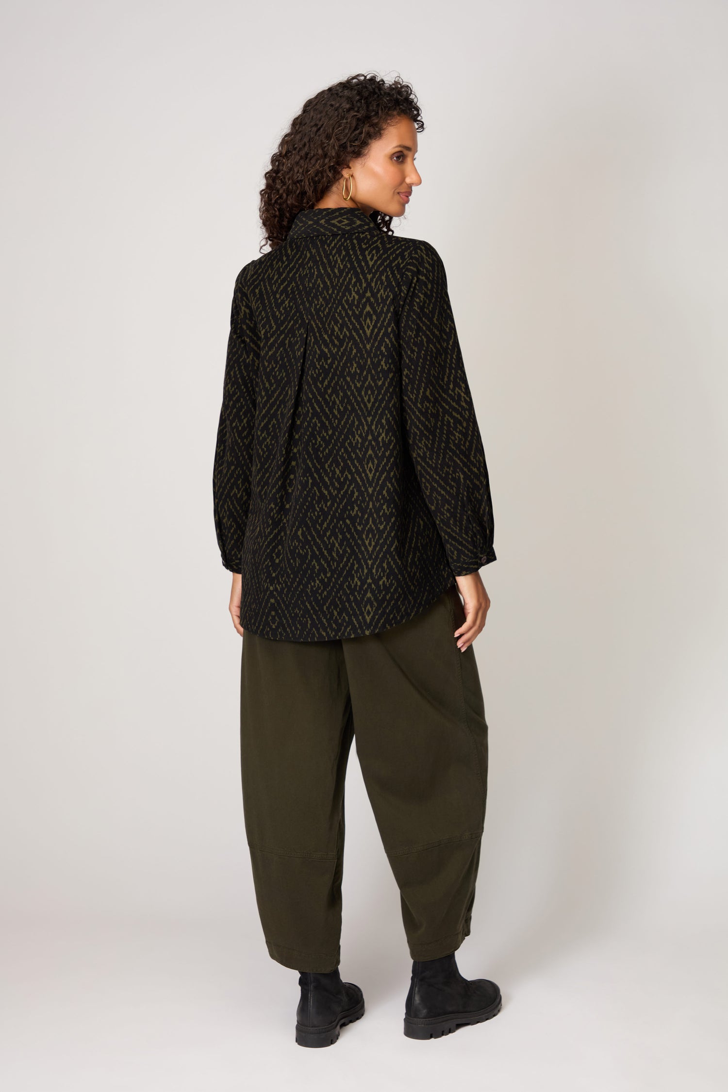 A person stands facing away in the Ornate Textured Cord Black and Khaki Shirt—an effortlessly stylish autumn wardrobe choice—paired with loose olive green pants and black boots against a plain light background.