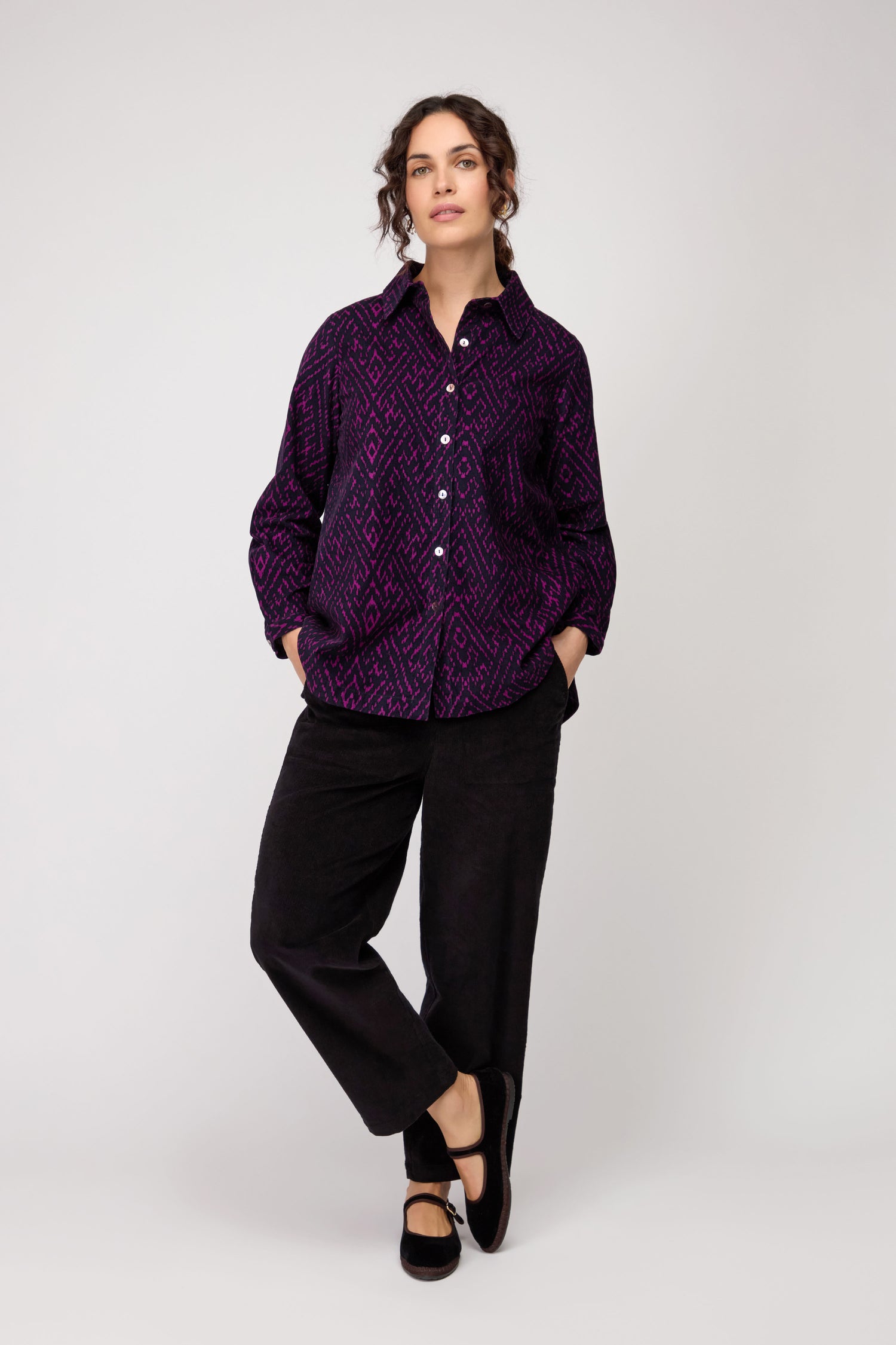 A person stands with hands in pockets, wearing the Ornate Textured Night and Plum Cord Shirt—featuring a relaxed fit and purple pattern—paired with black pants and shoes against a plain light background.
