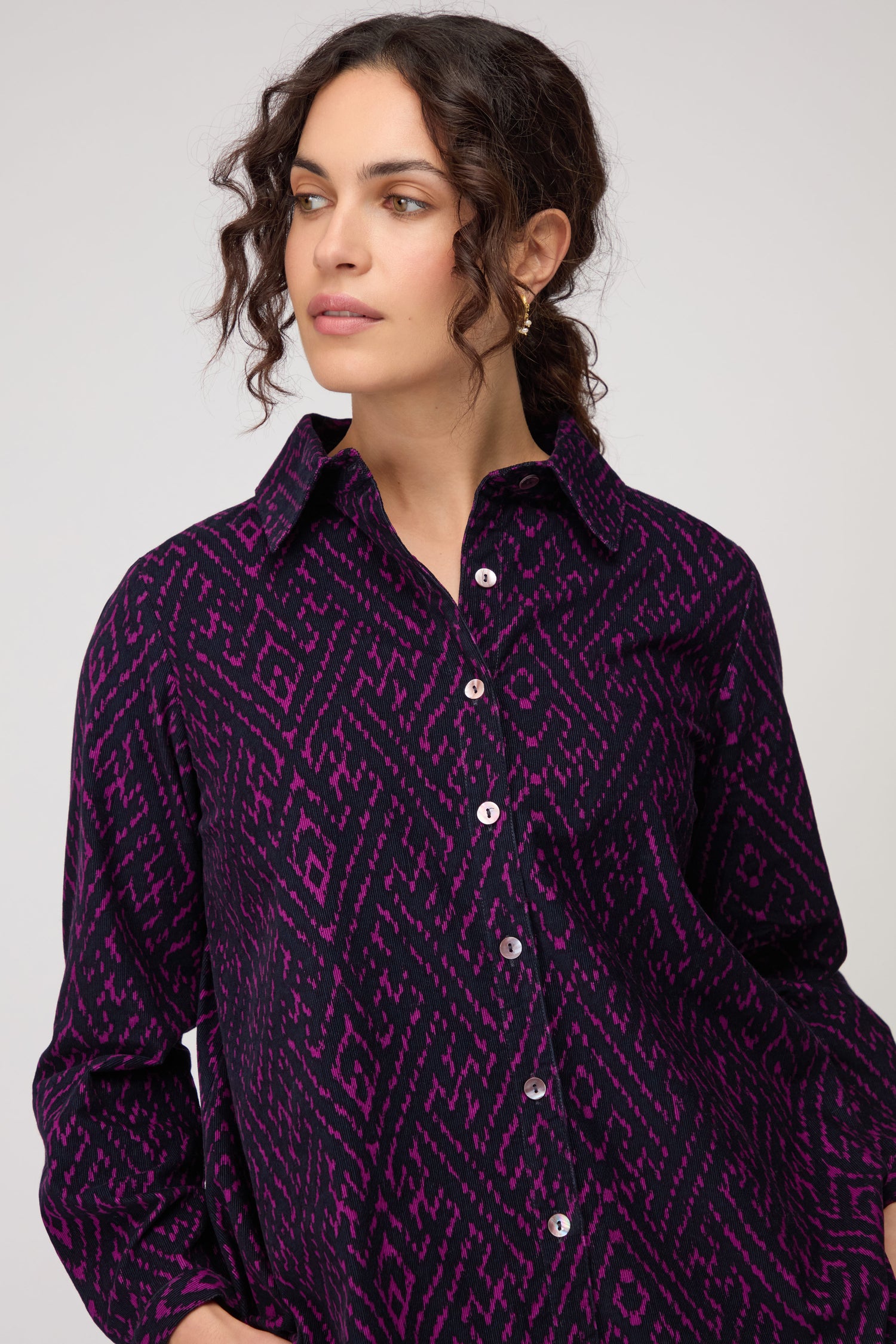 Woman with curly hair wears the Ornate Textured Night and Plum Cord Shirt, featuring a relaxed fit and purple-black pattern, as she looks slightly to the side against a plain background.