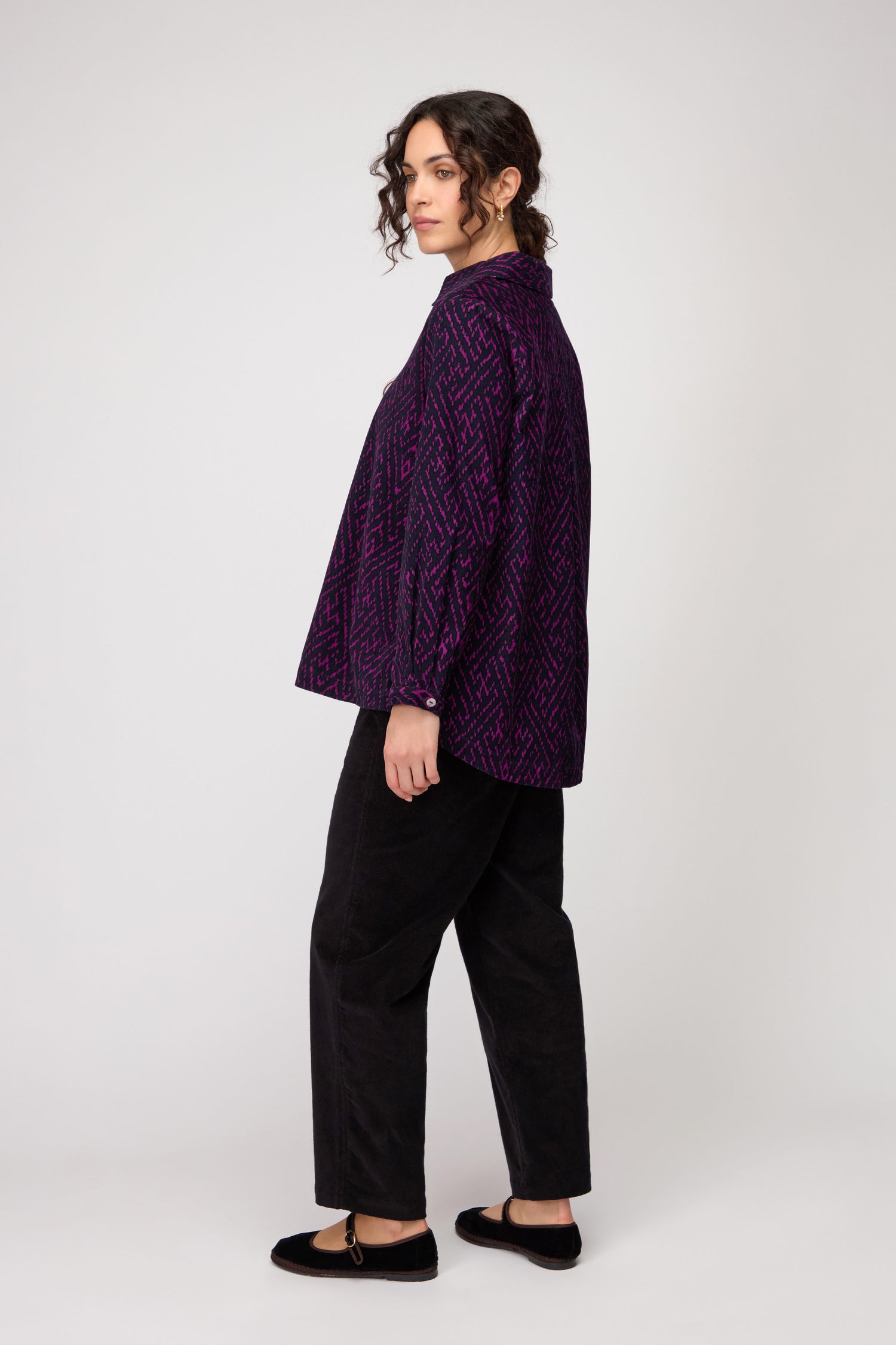 A person stands sideways, wearing the Ornate Textured Night and Plum Cord Shirt in a relaxed cut, paired with black pants and flats, set against a plain white background.