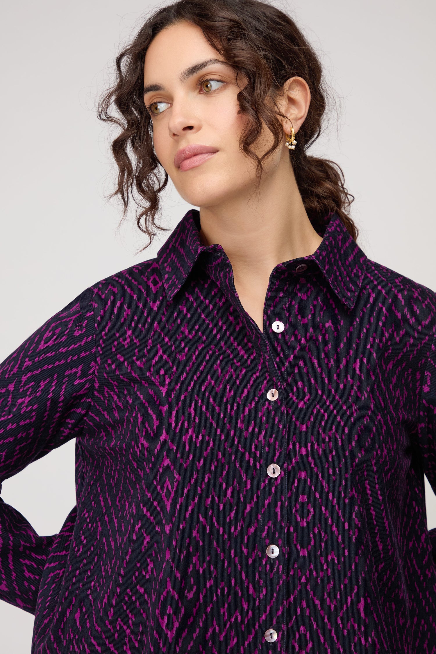 A woman with curly dark hair wears the Ornate Textured Night and Plum Cord Shirt, featuring a relaxed fit and geometric purple and black pattern, looking slightly to the side.