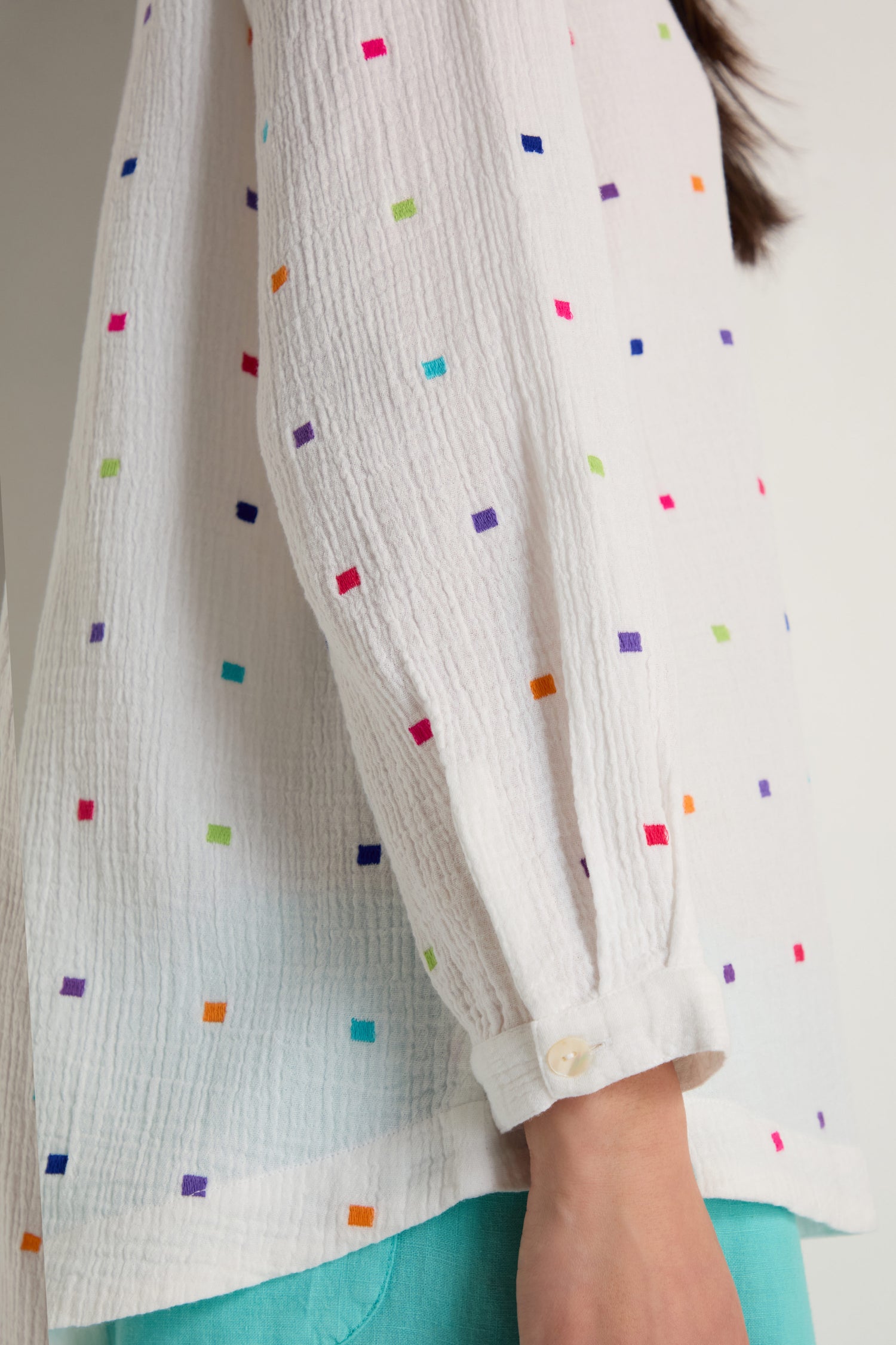 A person wears the Summer Squares Embroidered Shirt—a white, relaxed fit, textured gauzy cotton top with multicolored square embroidery and buttoned cuffs—paired with teal pants.