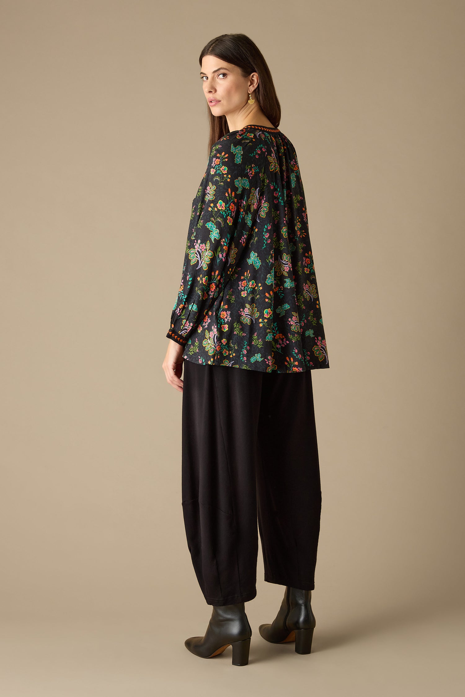 A woman wearing the Hand Drawn Floral Boxy Shirt paired with black pants stands in profile, gazing at the camera. Her chic look is finished with black boots, set against a simple beige backdrop.