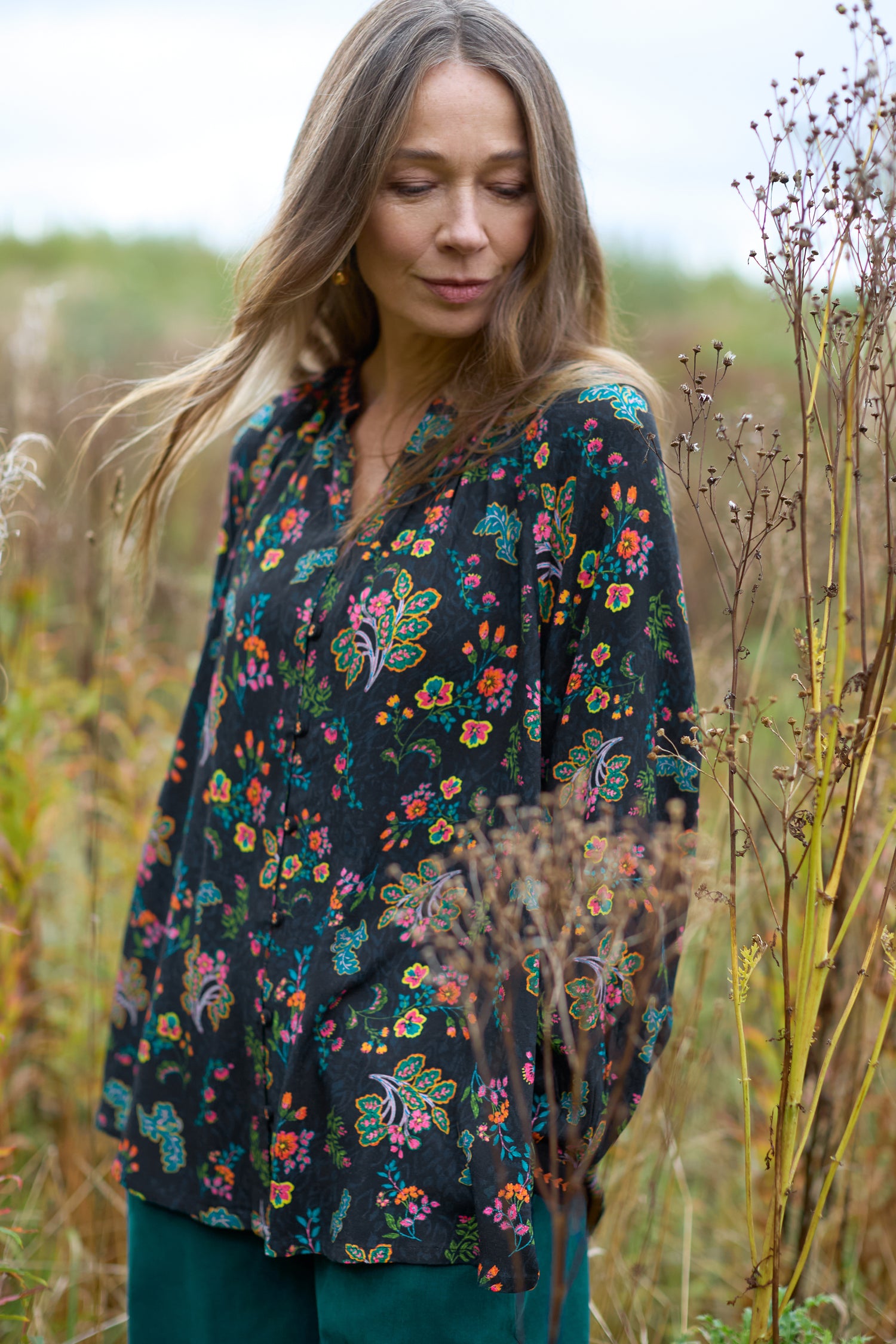 A woman wearing a Hand Drawn Floral Boxy Shirt is standing in a field surrounded by tall plants. Her eyes are closed, and the wind tousles her hair, enhancing the flowing silhouette of her attire.