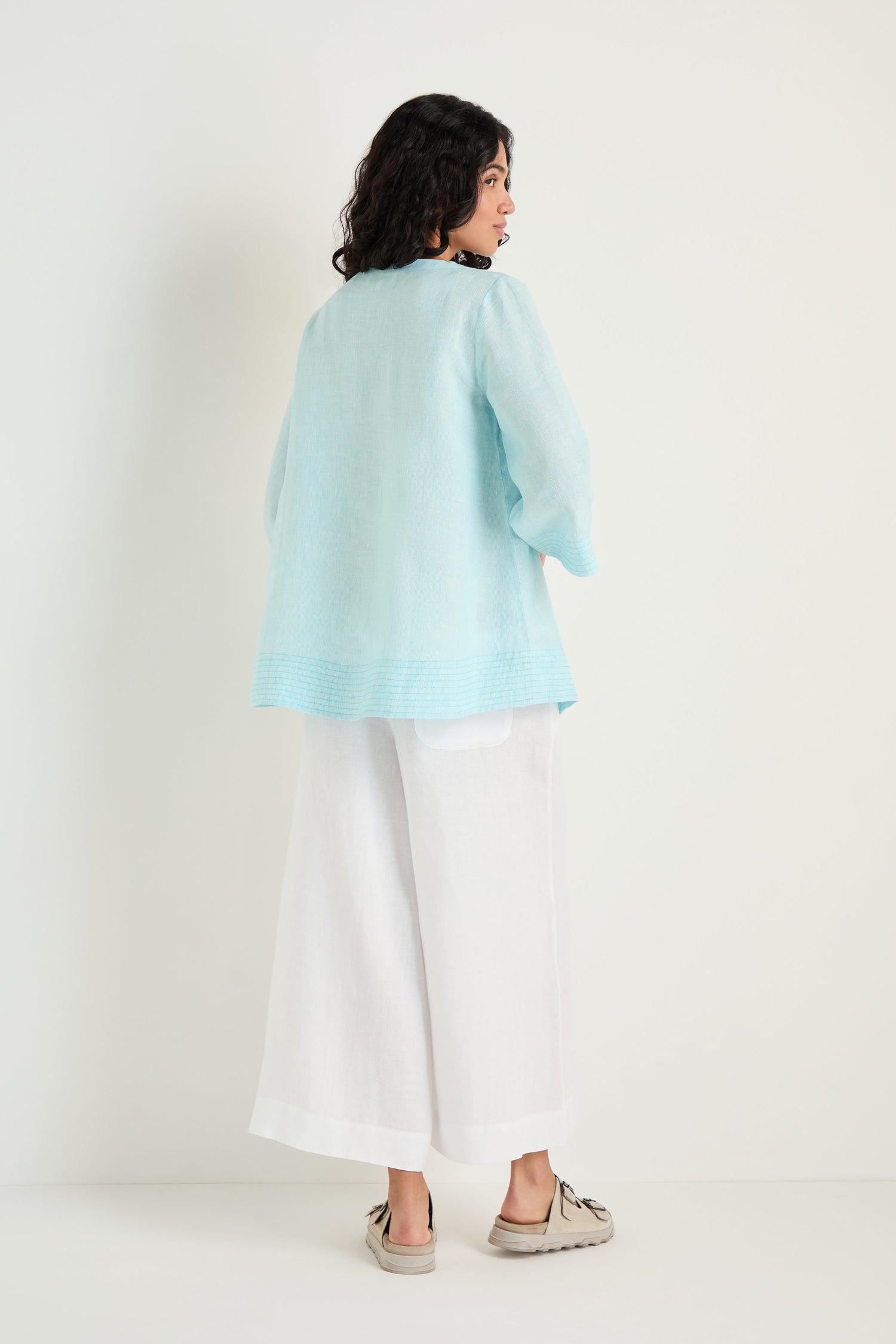 A person stands facing away, wearing the Ticking Stripe Linen Button Placket Tunic in light blue with an A-line silhouette, wide white pants, and beige shoes against a plain white background.