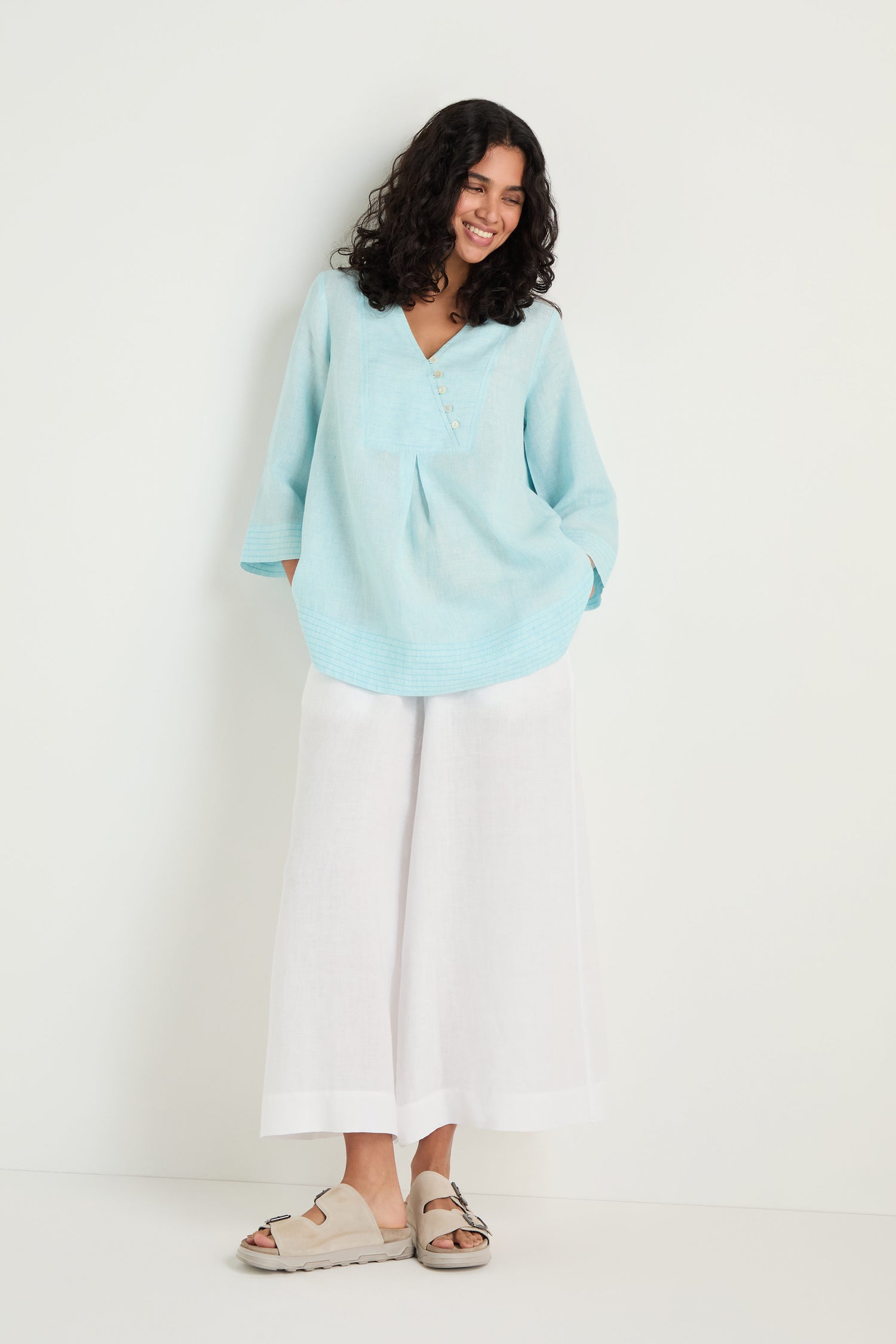 A woman with curly hair stands against a white wall, hands in pockets, wearing the Ticking Stripe Linen Button Placket Tunic in light blue stripes, paired with wide white pants and beige sandals.