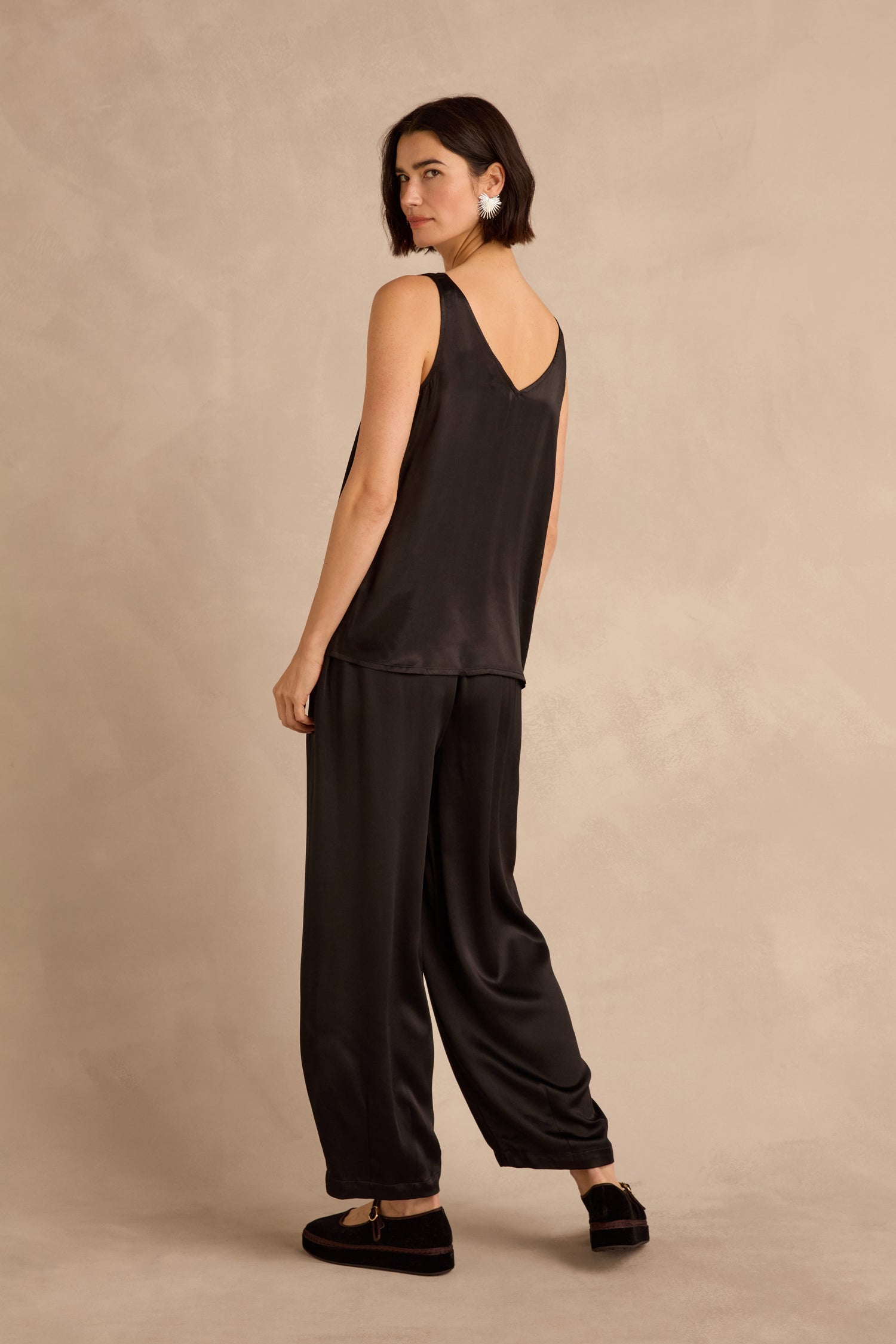 Woman wearing the Cupro Layering Vest and loose black pants stands on a neutral background, looking over her shoulder.