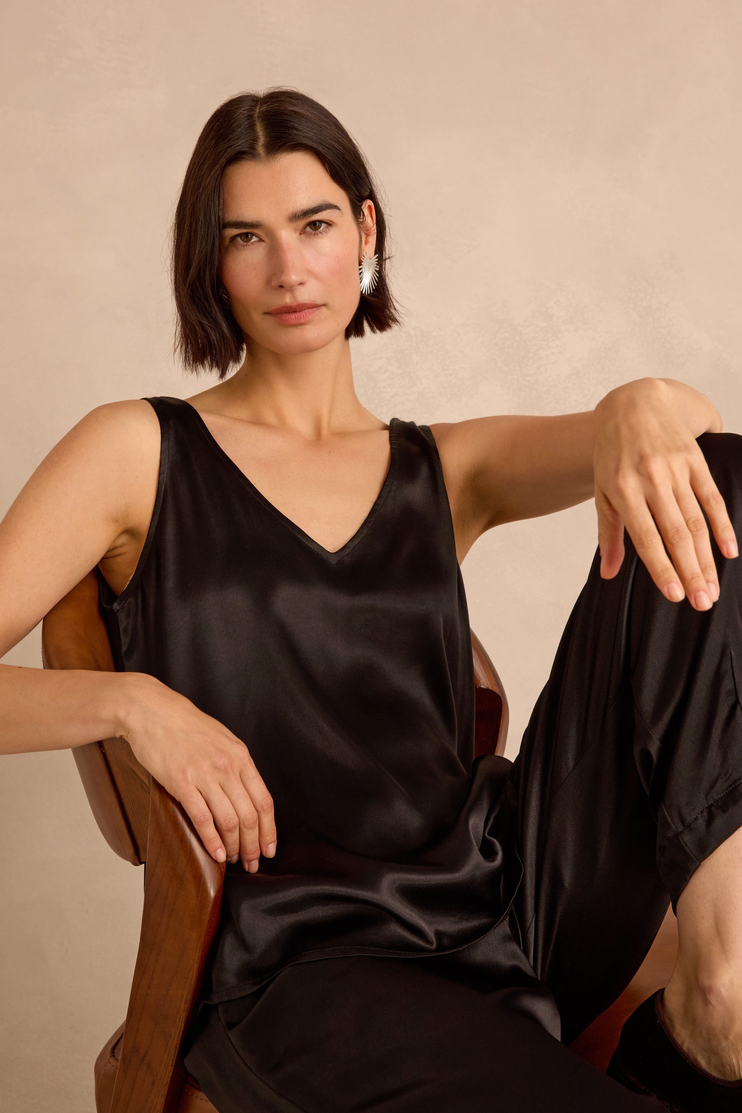 A woman with straight brown hair sits in a wooden chair, wearing the black Cupro Layering Vest and black pants, looking directly at the camera with a neutral expression.